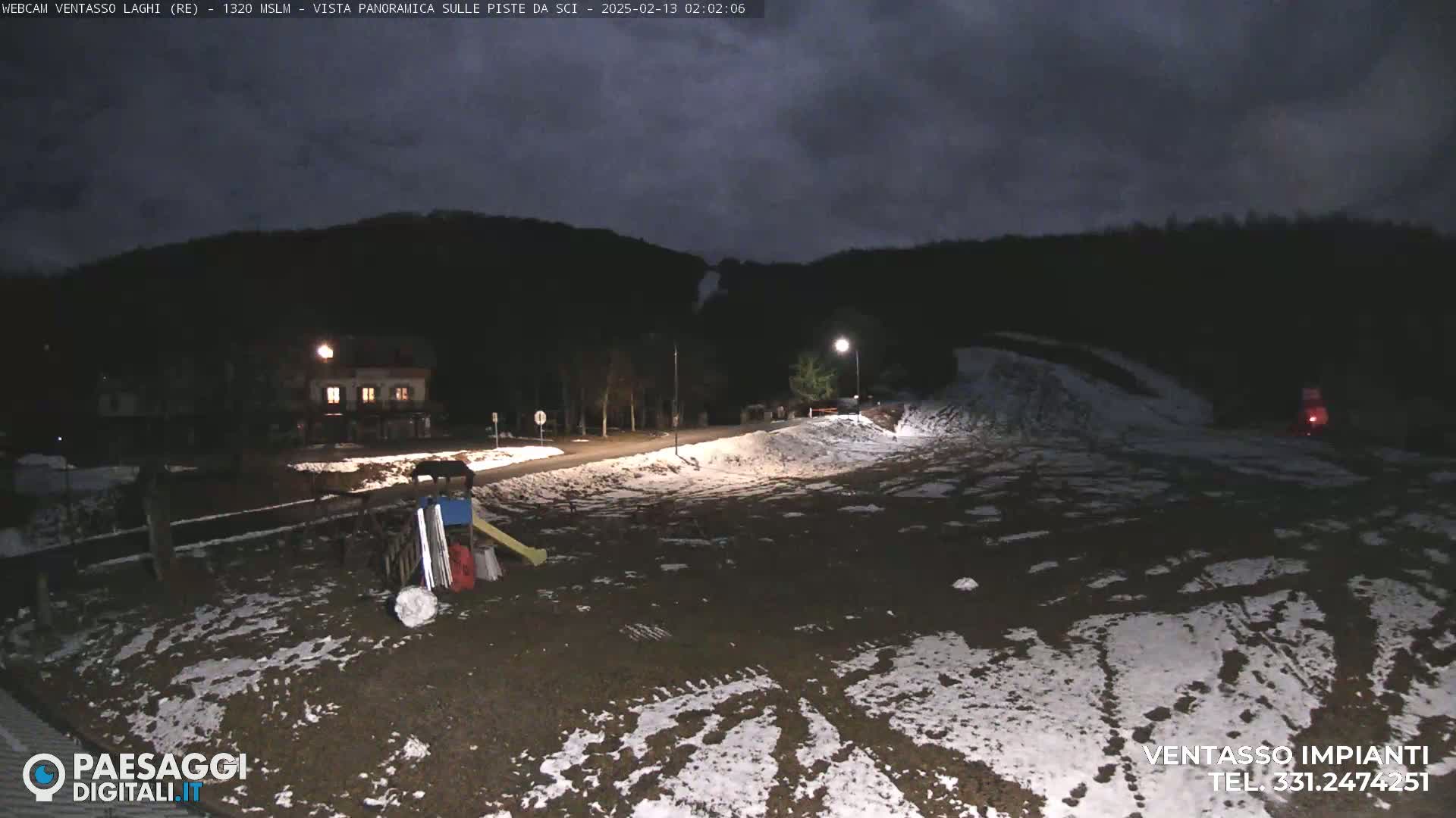 A nighttime view of a snow-dusted, sparsely populated area with a building, a road, and playground equipment under a cloudy sky.