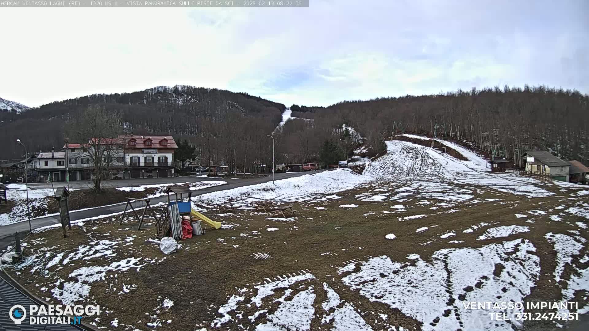 A partially snow-covered playground and ski slope are visible in front of a village nestled in a mountainous area under an overcast sky.