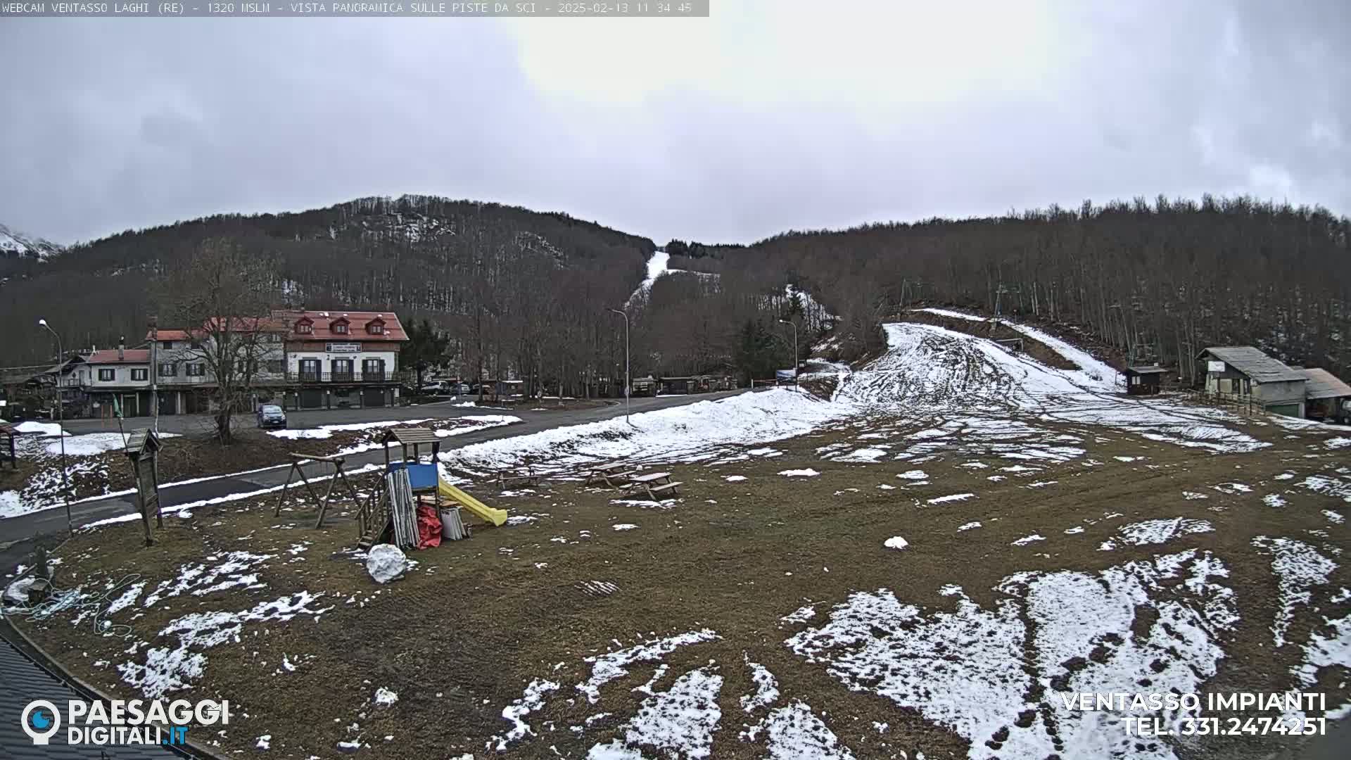 A mostly cloudy day reveals a snow-dusted ski slope and playground near a village nestled at the base of a wooded hill.