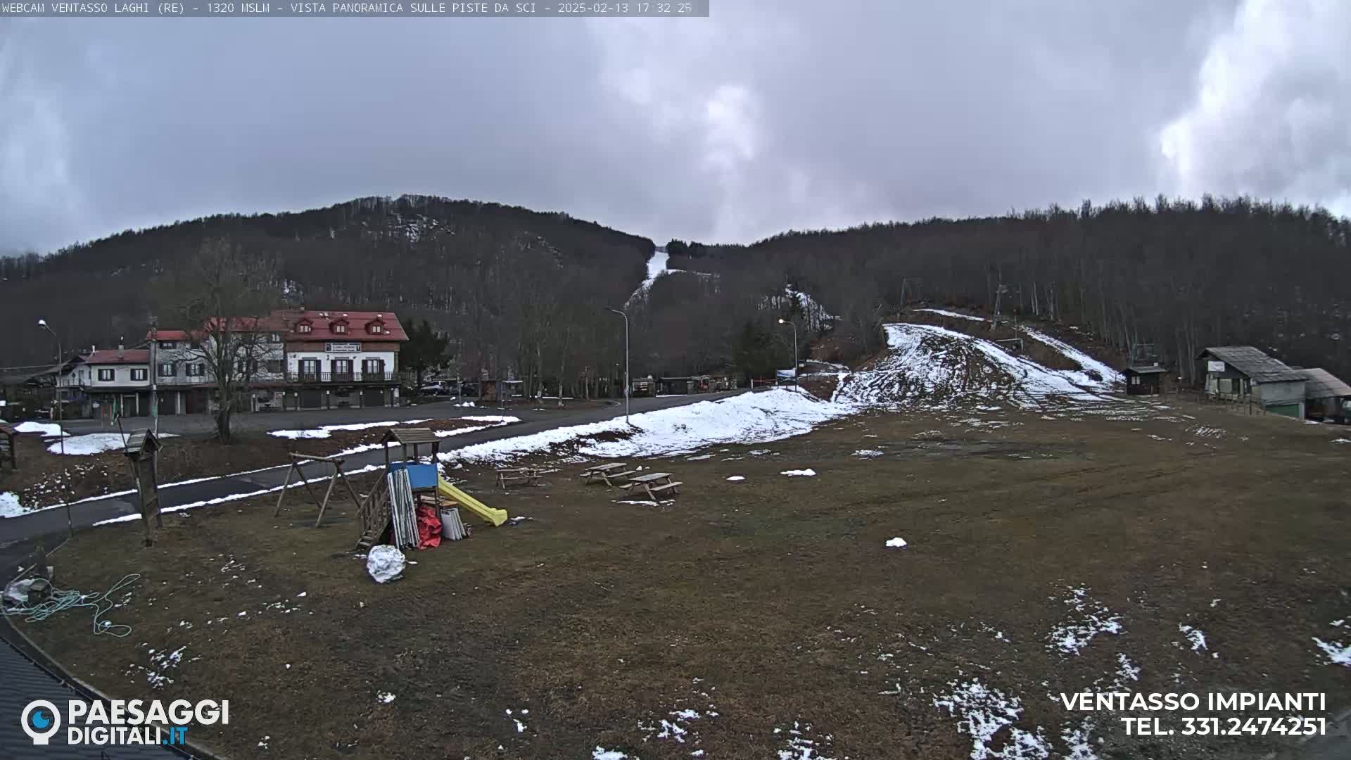 A mostly cloudy day reveals a ski slope with patches of snow, a playground, several buildings, and a sparsely vegetated area in the foreground.