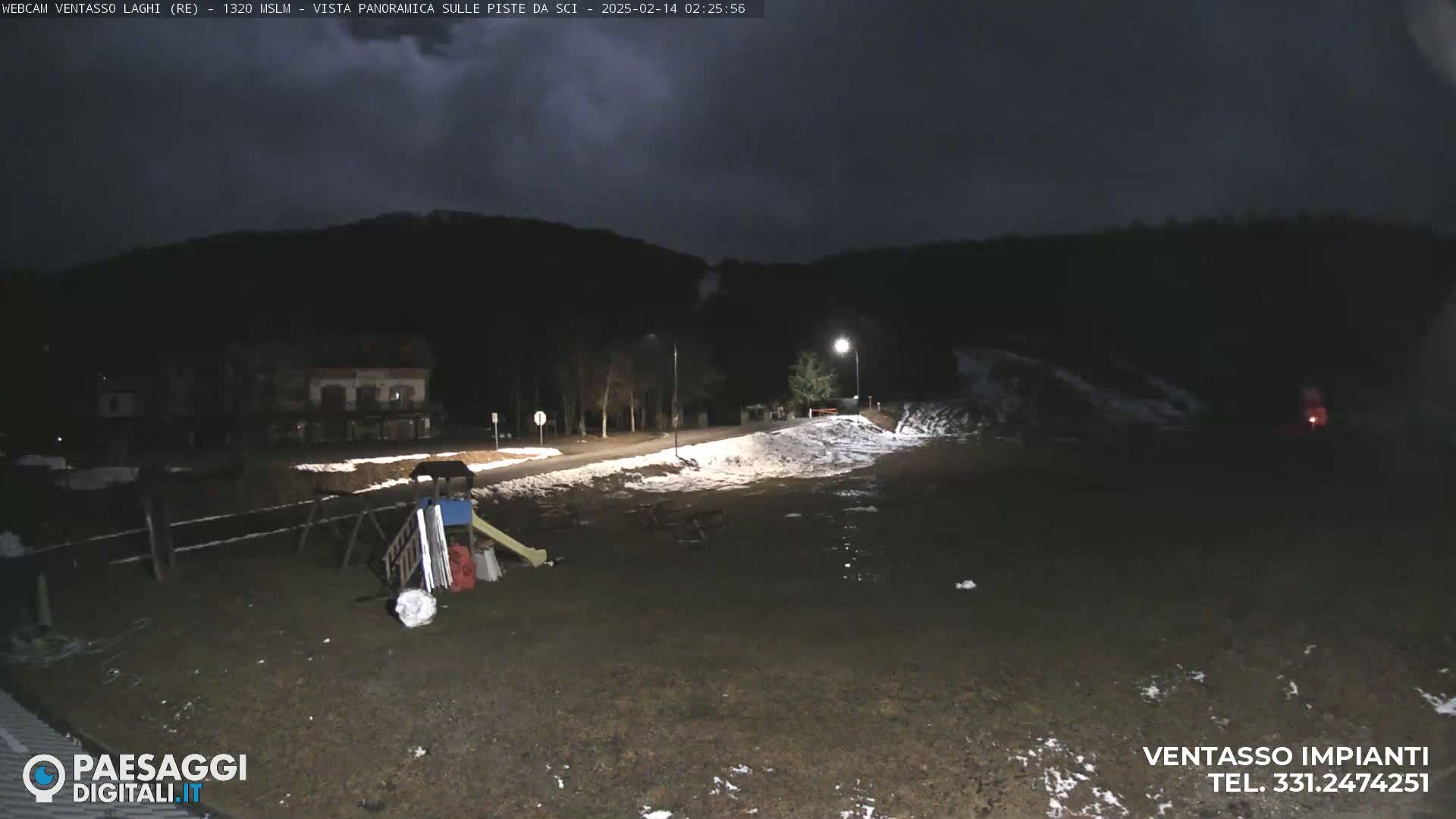 A nighttime view of a playground with some snow on the ground, near a road and dark forested hills under a cloudy sky.