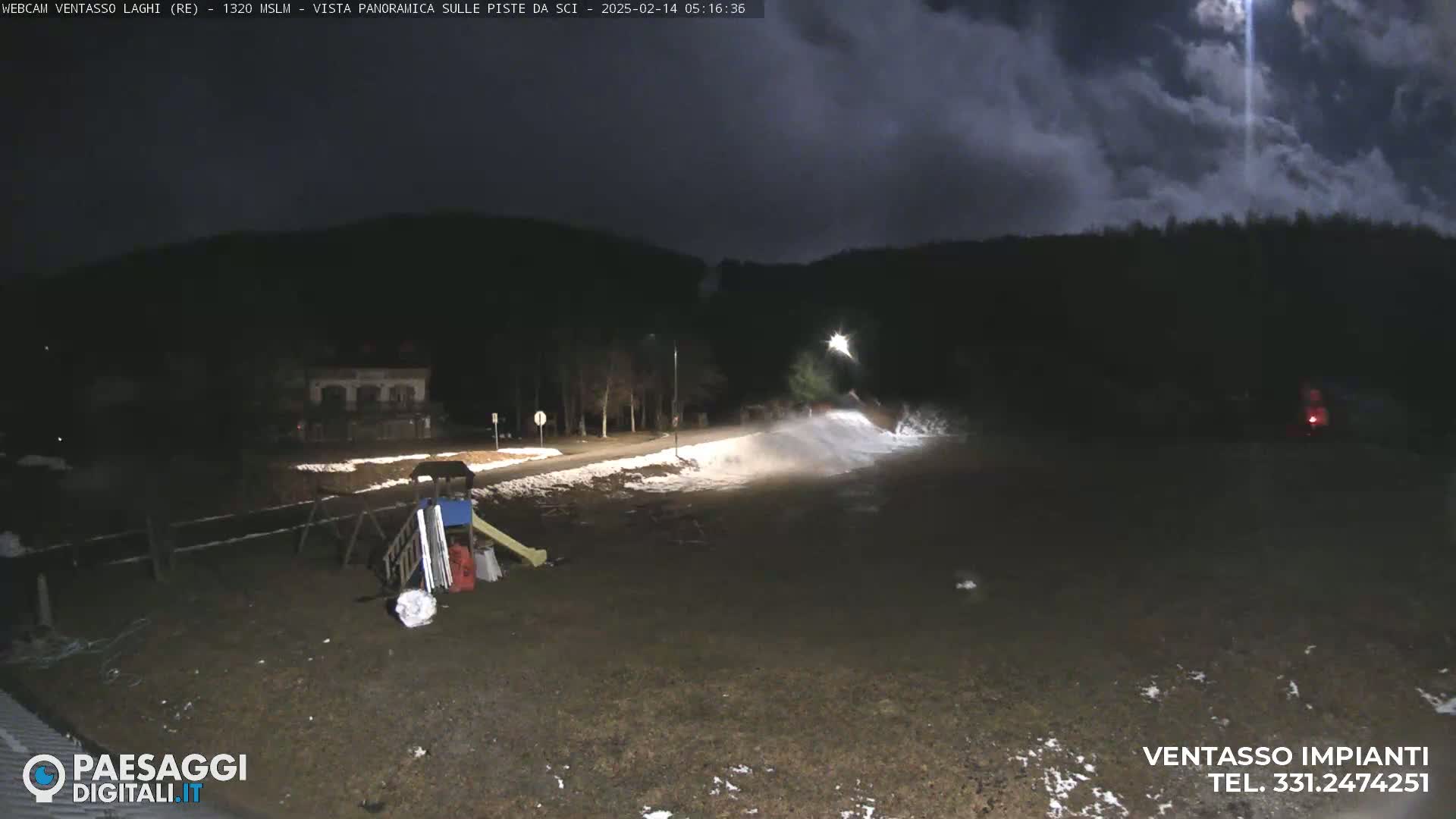 A nighttime view of a snow-covered area with a playground, a building in the distance, and a dark, cloudy sky.