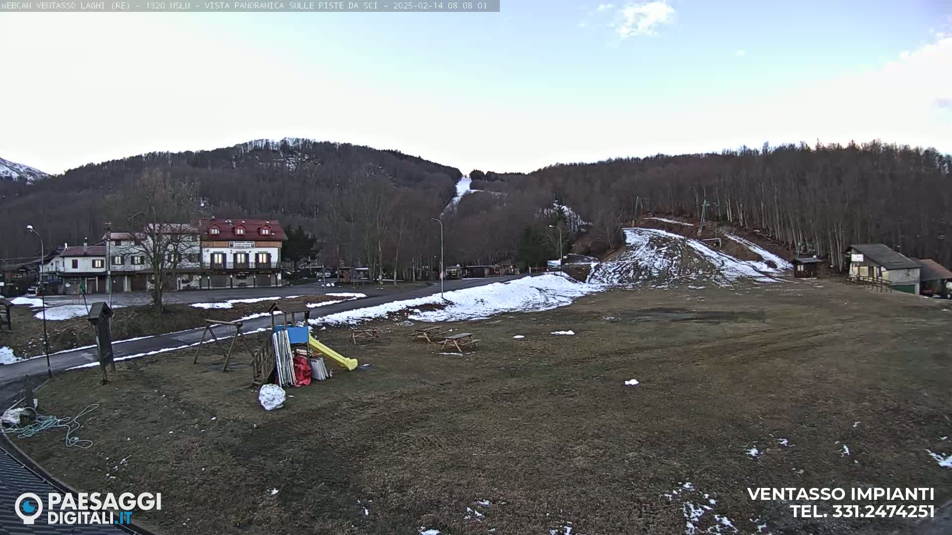 A playground with a slide and swings sits in front of a small building near a ski slope with patchy snow, under a mostly clear sky.