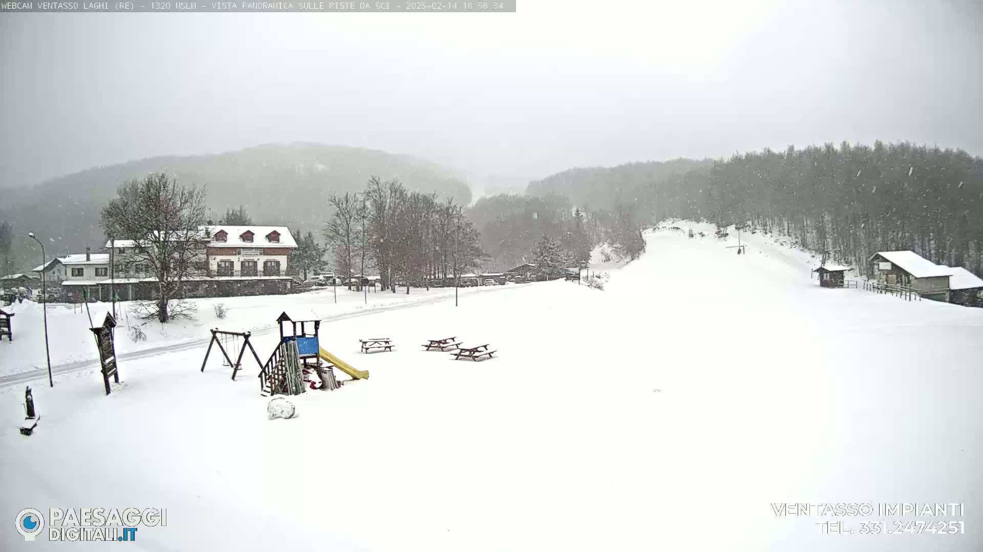 A snow-covered landscape with buildings, a playground, and picnic tables, under a lightly snowing sky.