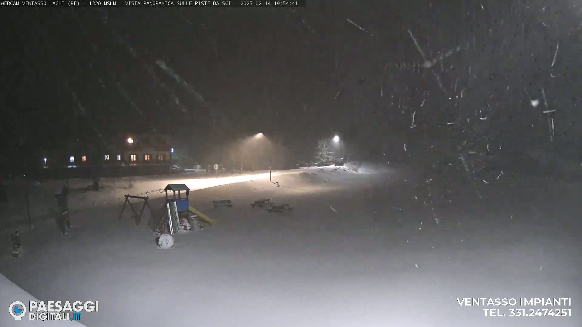 A snow-covered playground and a road are illuminated by streetlights under a heavy snowfall at night.