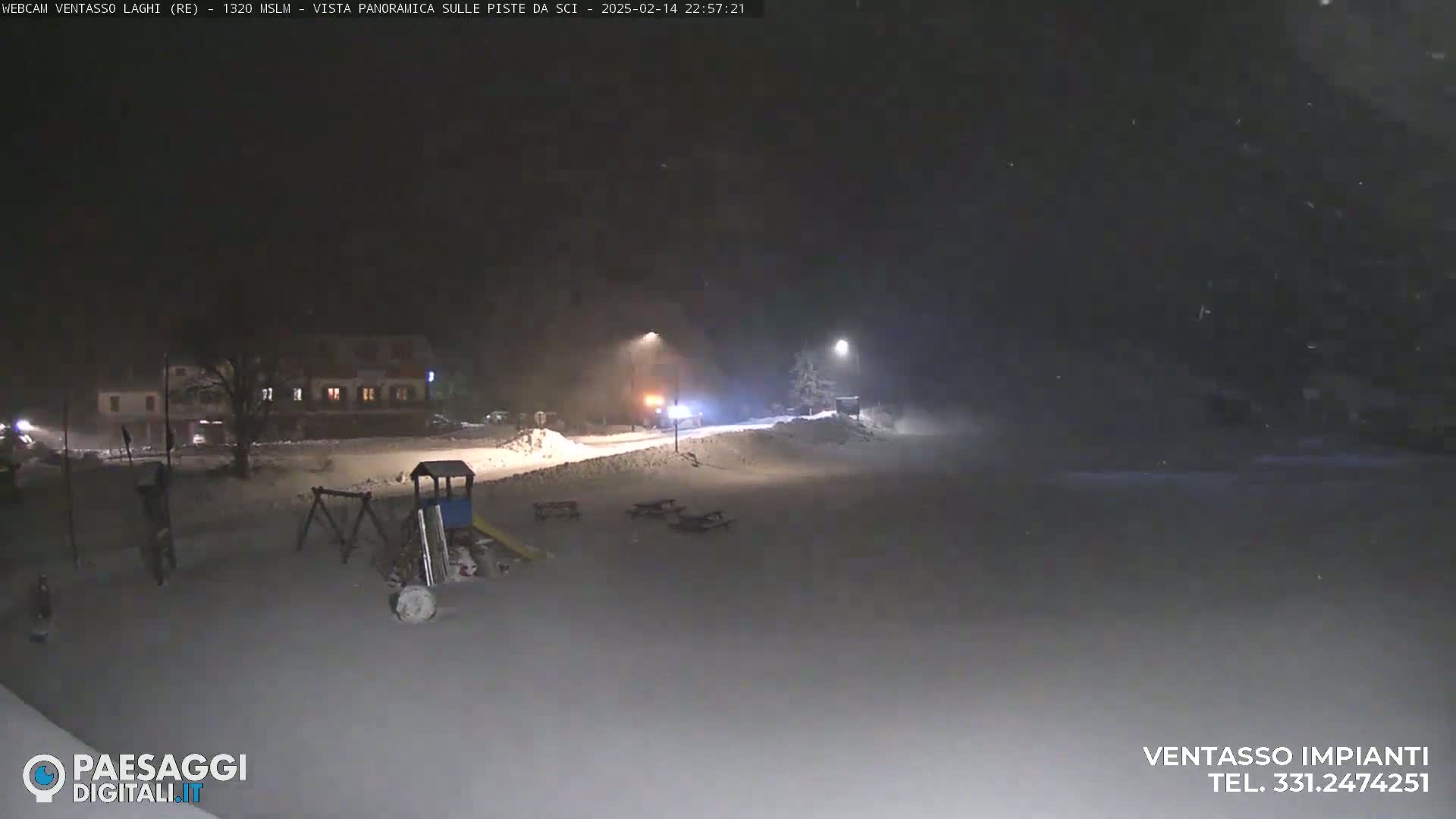 A snow-covered playground and picnic area are illuminated at night under falling snow near a building.