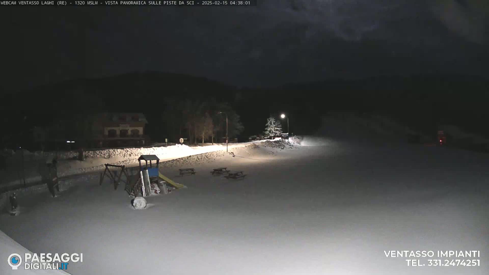 A snow-covered playground with picnic tables is illuminated at night near a building and dark trees under a cloudy sky.