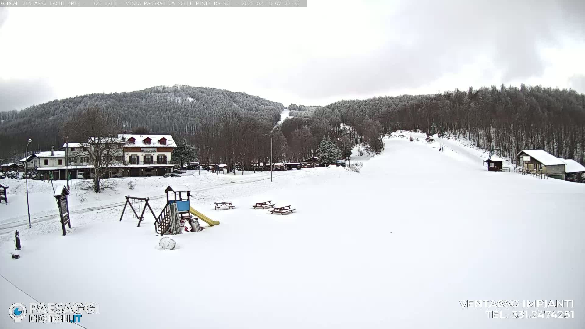 A snow-covered playground with picnic tables sits in front of a building and a ski slope, all nestled amongst snow-covered trees under an overcast sky.