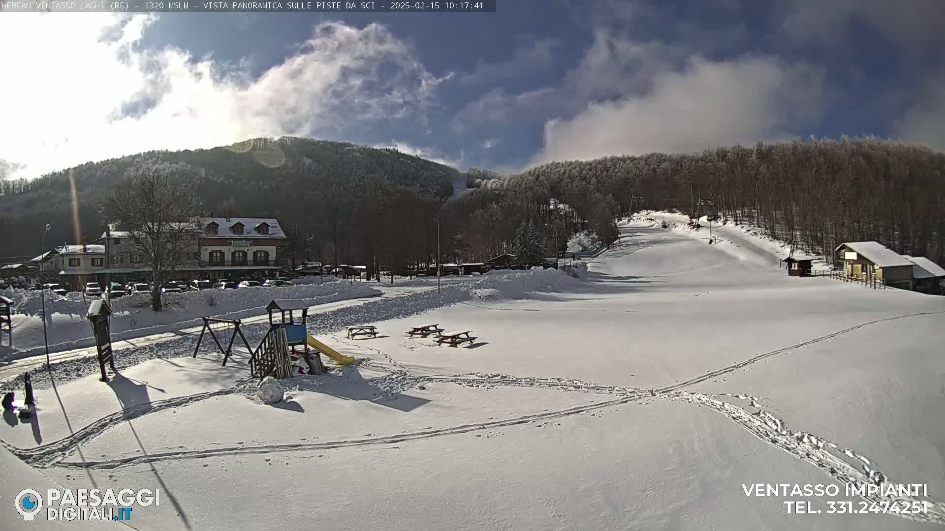 A snow-covered ski resort scene under a partly sunny sky shows a playground, picnic tables, a building, and a ski slope with tracks in the snow.