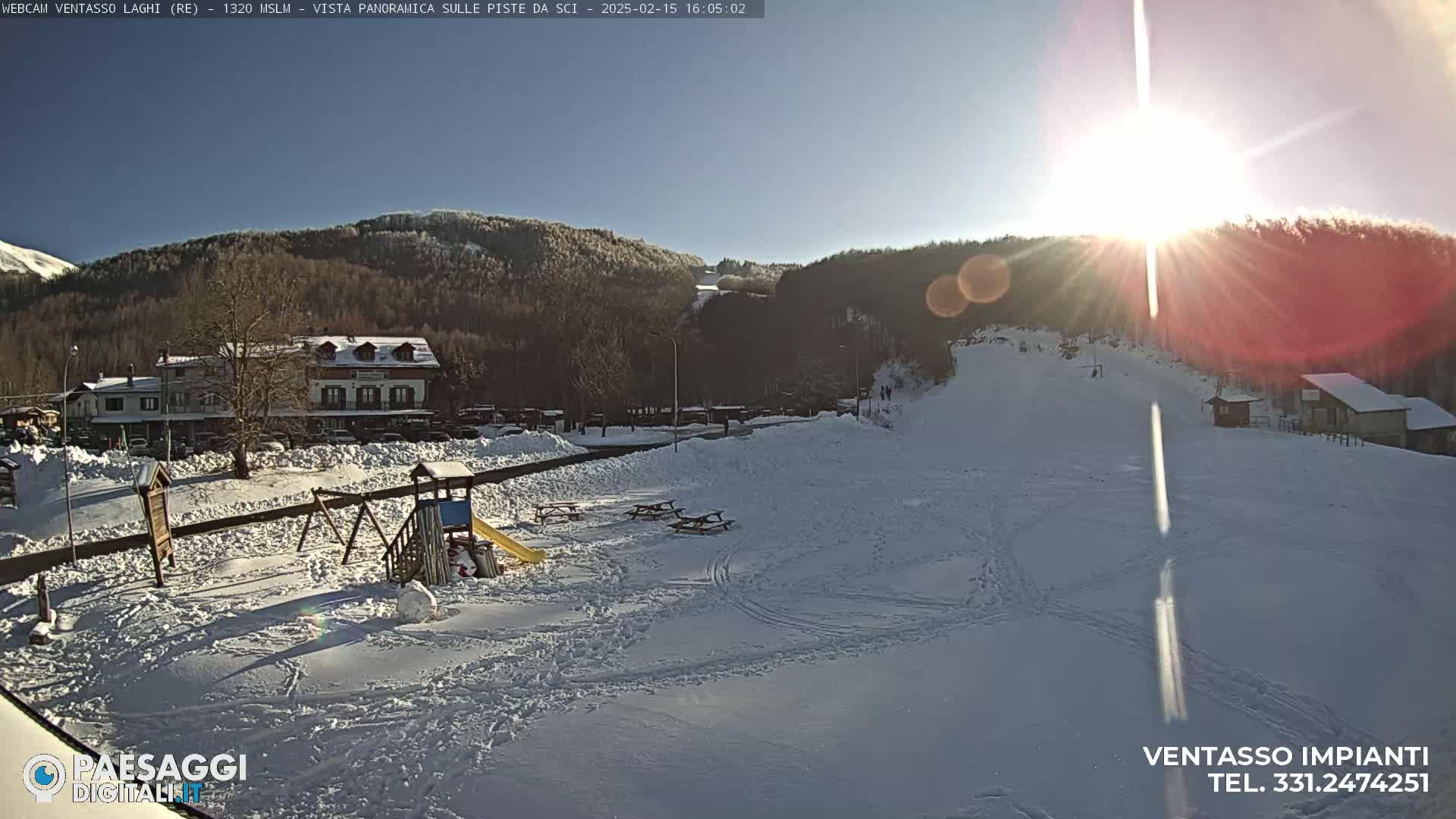A sunny, snow-covered landscape features a playground, picnic tables, buildings, and a ski slope, all set against a backdrop of hills and mountains.