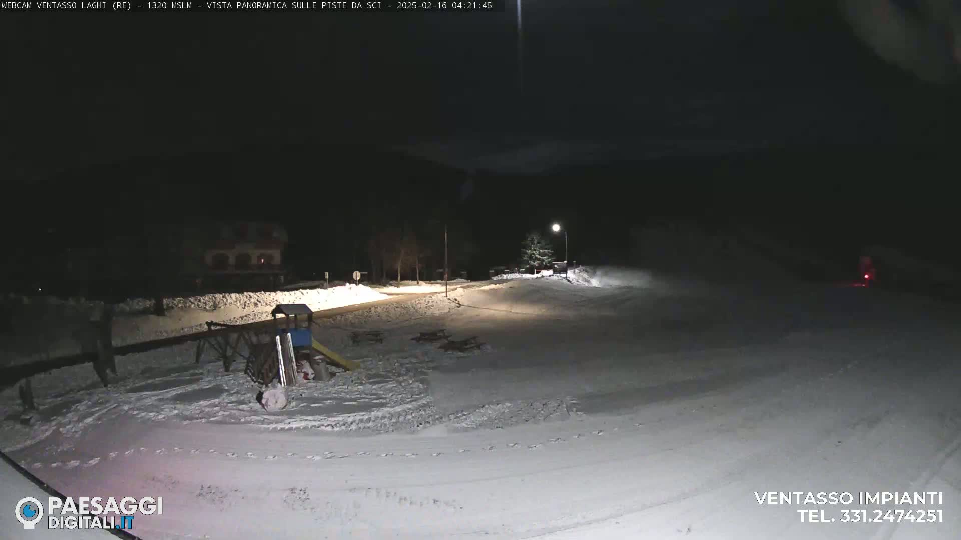 A snow-covered area at night, illuminated by a streetlight, shows a playground and a building in the distance.