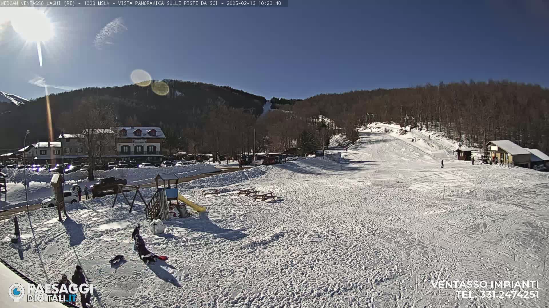 A sunny day at a snow-covered ski resort, showing a playground, buildings, a ski slope, and several people.