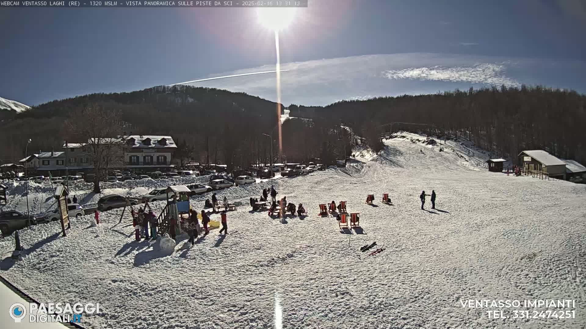 A sunny day at a snow-covered ski resort shows a parking lot full of cars, people gathered in a snow-covered area with deck chairs, a playground, and a ski slope in the background.