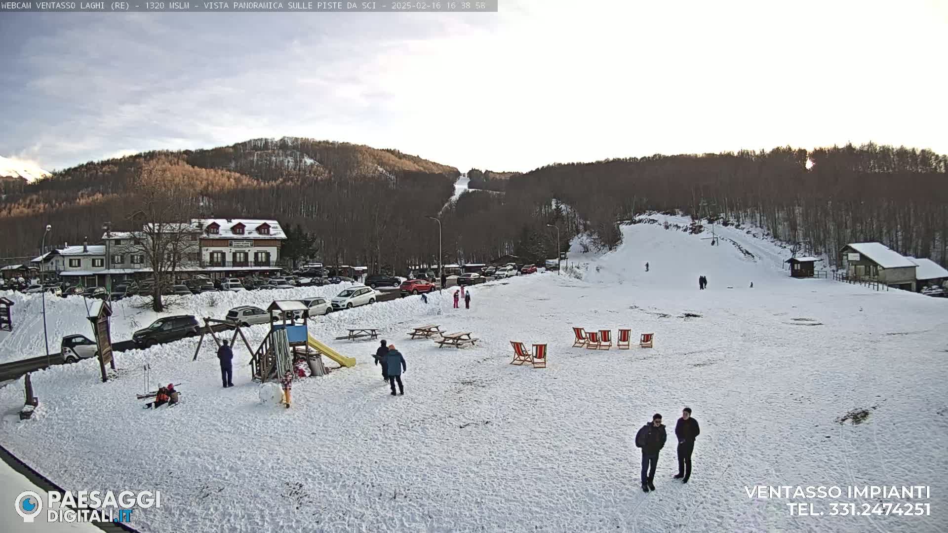 A snow-covered parking lot with a playground and several people, situated below a ski slope and a wooded hillside under a partly cloudy sky.