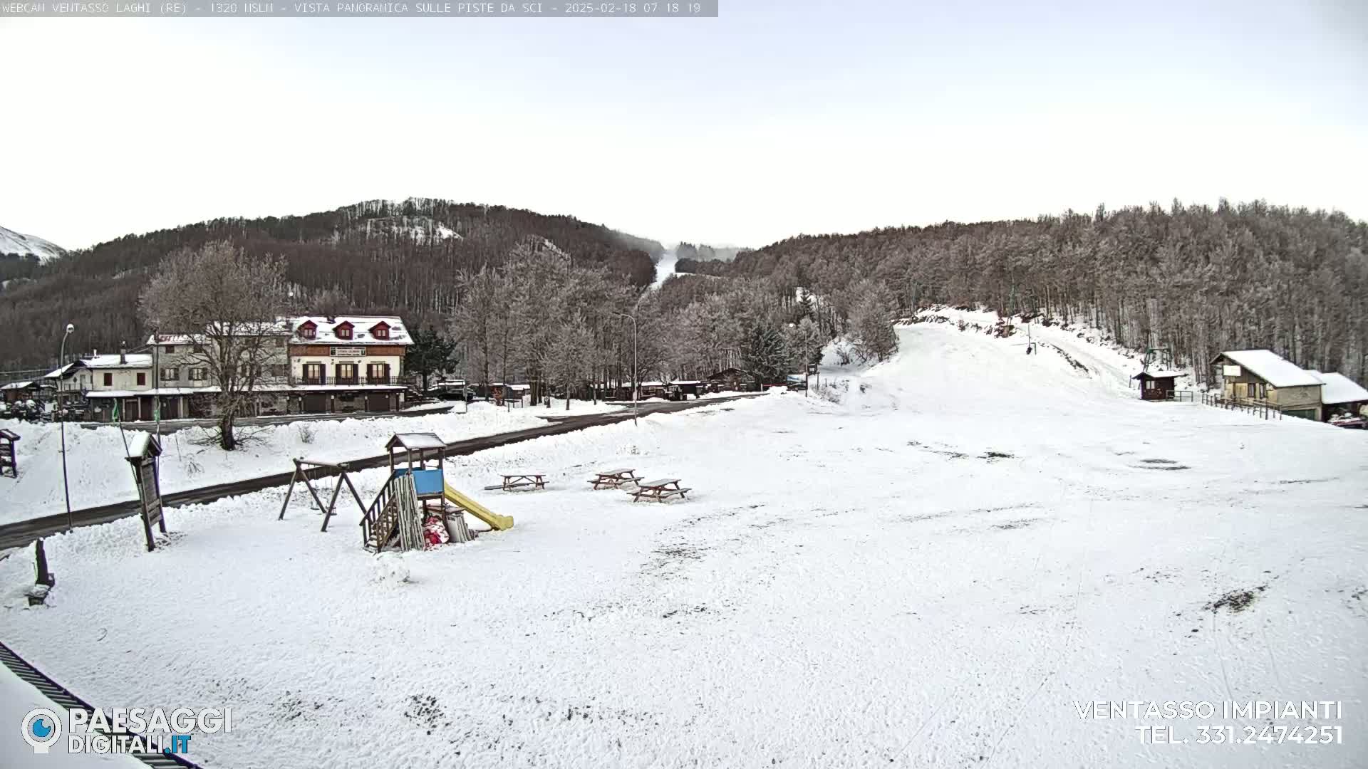 A snow-covered playground and picnic area sit in front of a ski slope, with a village and snow-covered hills in the background under a cloudy sky.