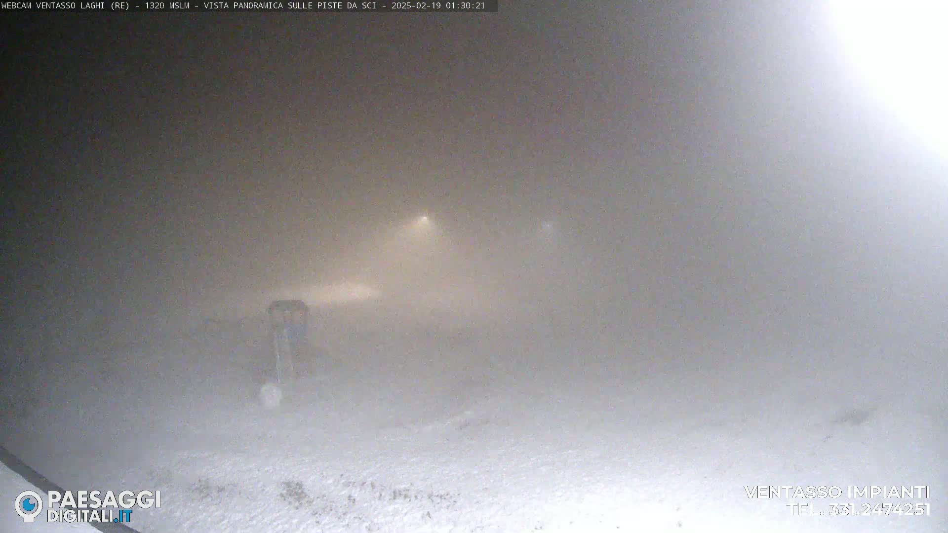 A snow-covered landscape is barely visible through a thick fog at night, illuminated by faint lights in the distance.