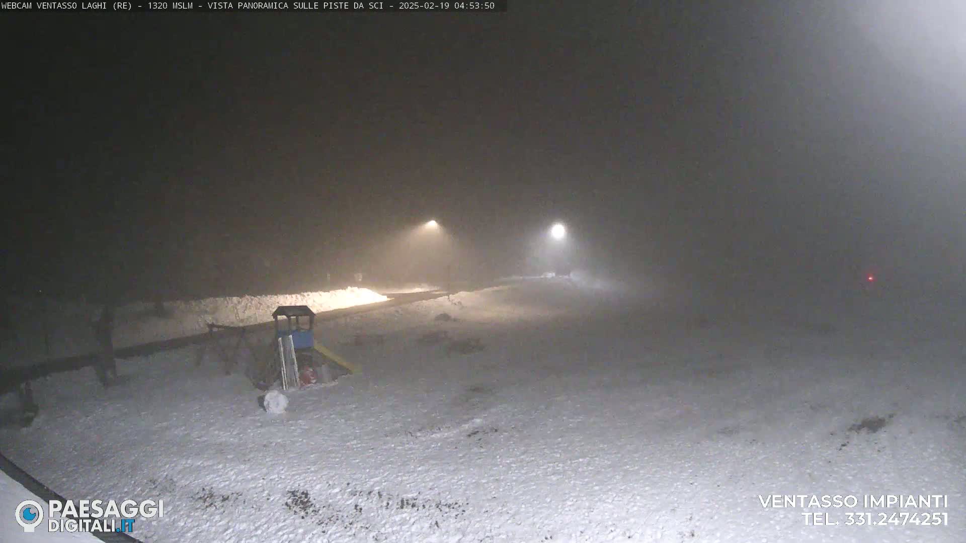 A snow-covered playground is dimly lit by two distant lights in foggy nighttime conditions.
