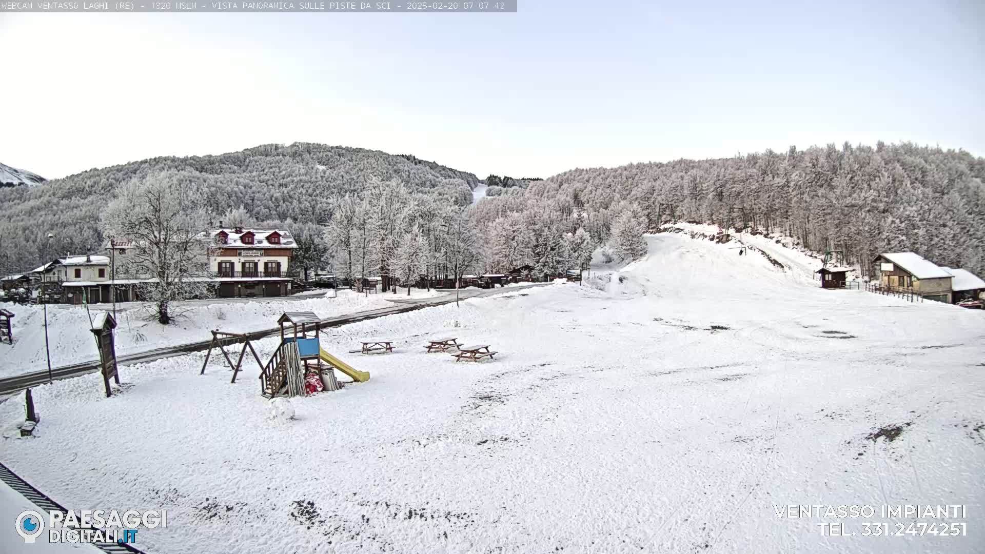 A snow-covered playground and small village are nestled at the base of a snow-dusted hill with a ski slope visible in the background under a clear, light-blue sky.