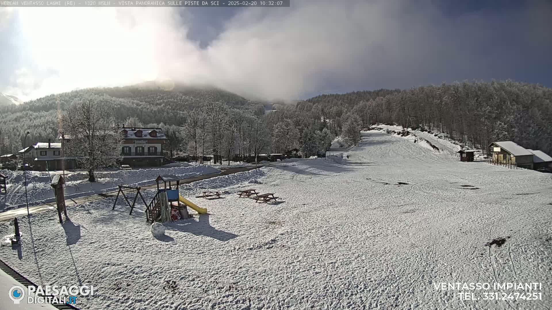 A snow-covered playground and ski slope are situated below a partly cloudy sky, with a village and snow-covered trees in the background.