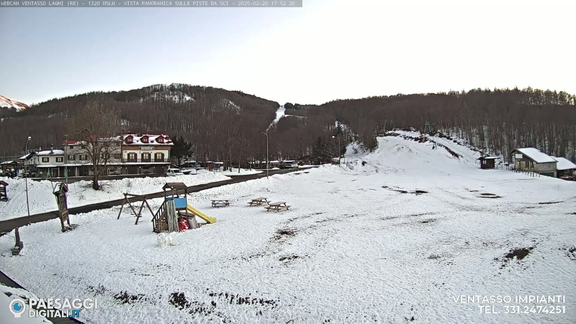 A snow-covered playground and picnic area sit in front of a building and ski slope in a wooded area.