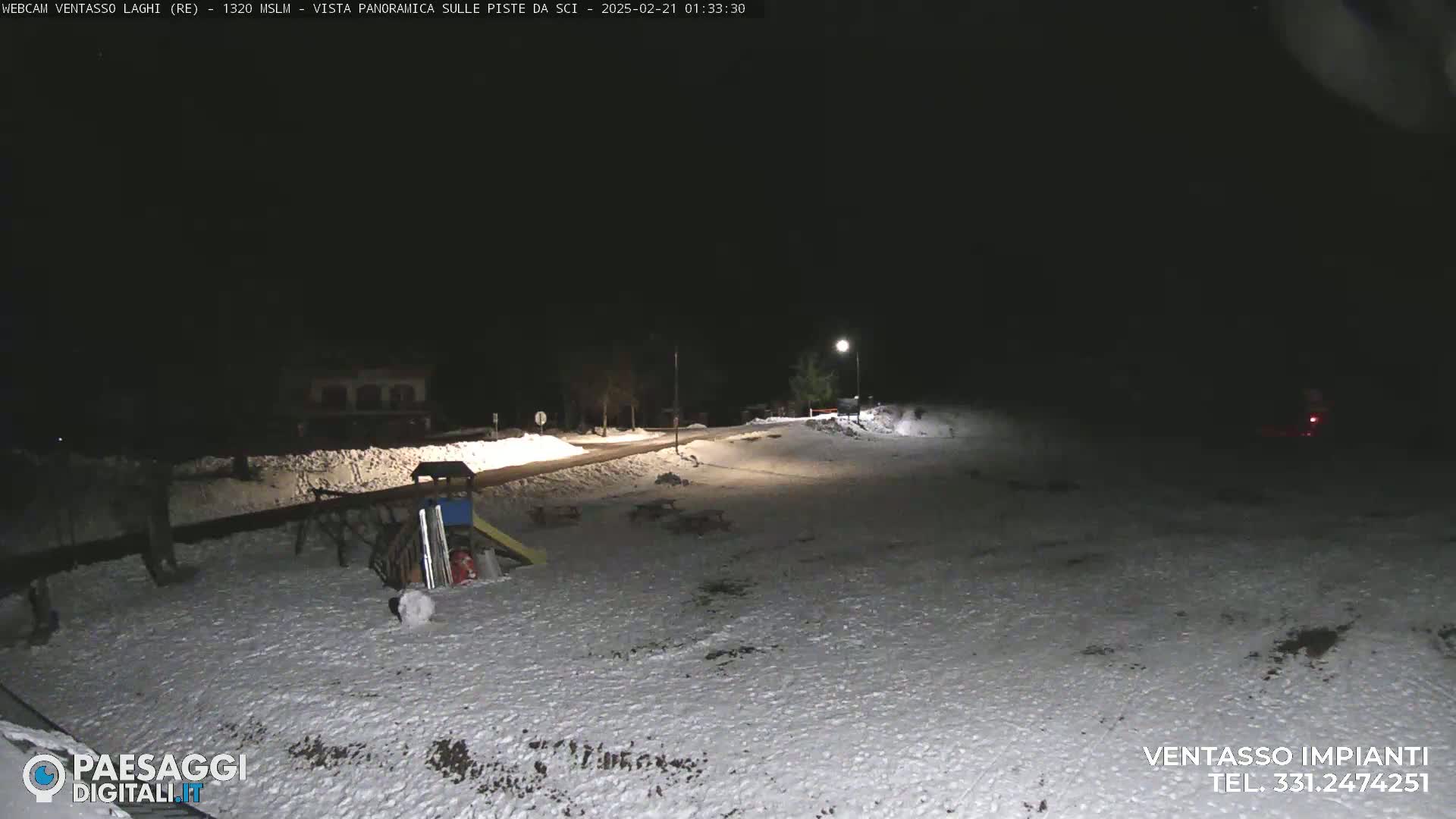 A snow-covered area at night, illuminated by artificial lights, shows a playground and a road, with a building visible in the distance.
