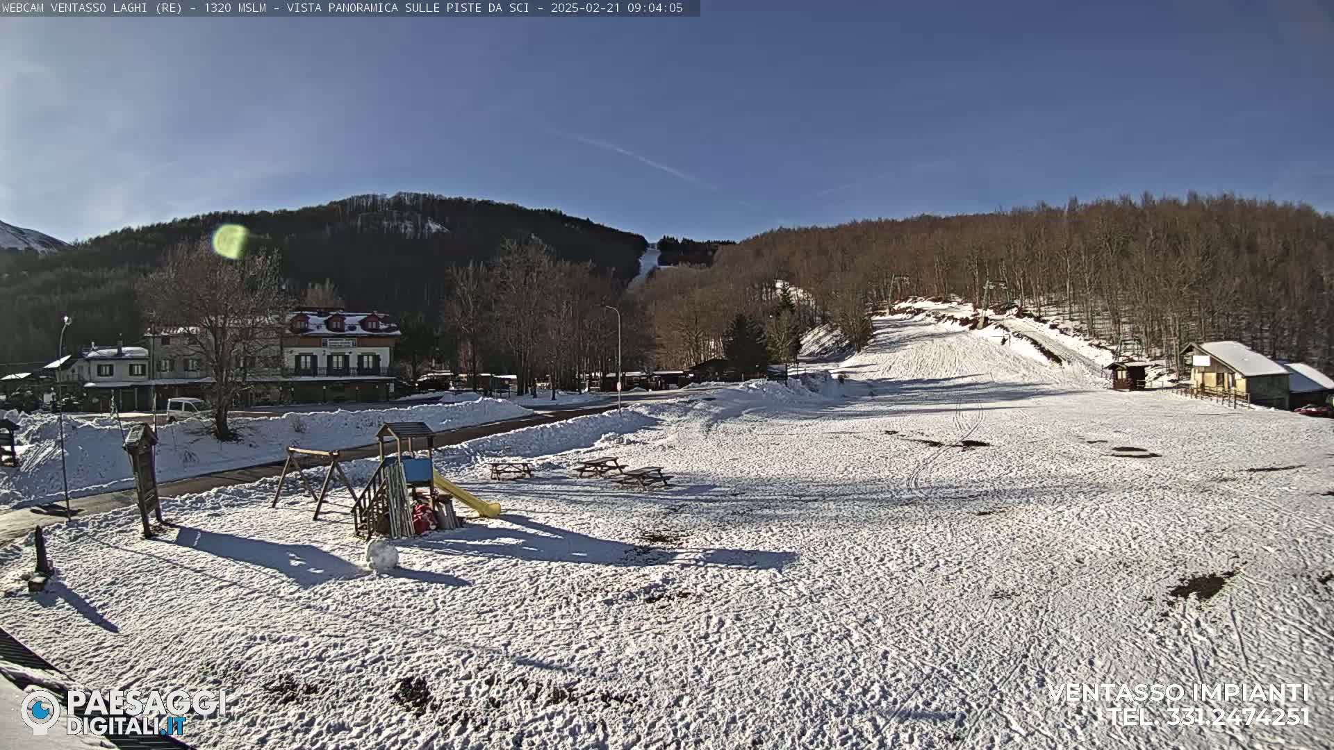 A snow-covered playground and ski slope are visible, situated below a line of leafless trees under a clear blue sky.