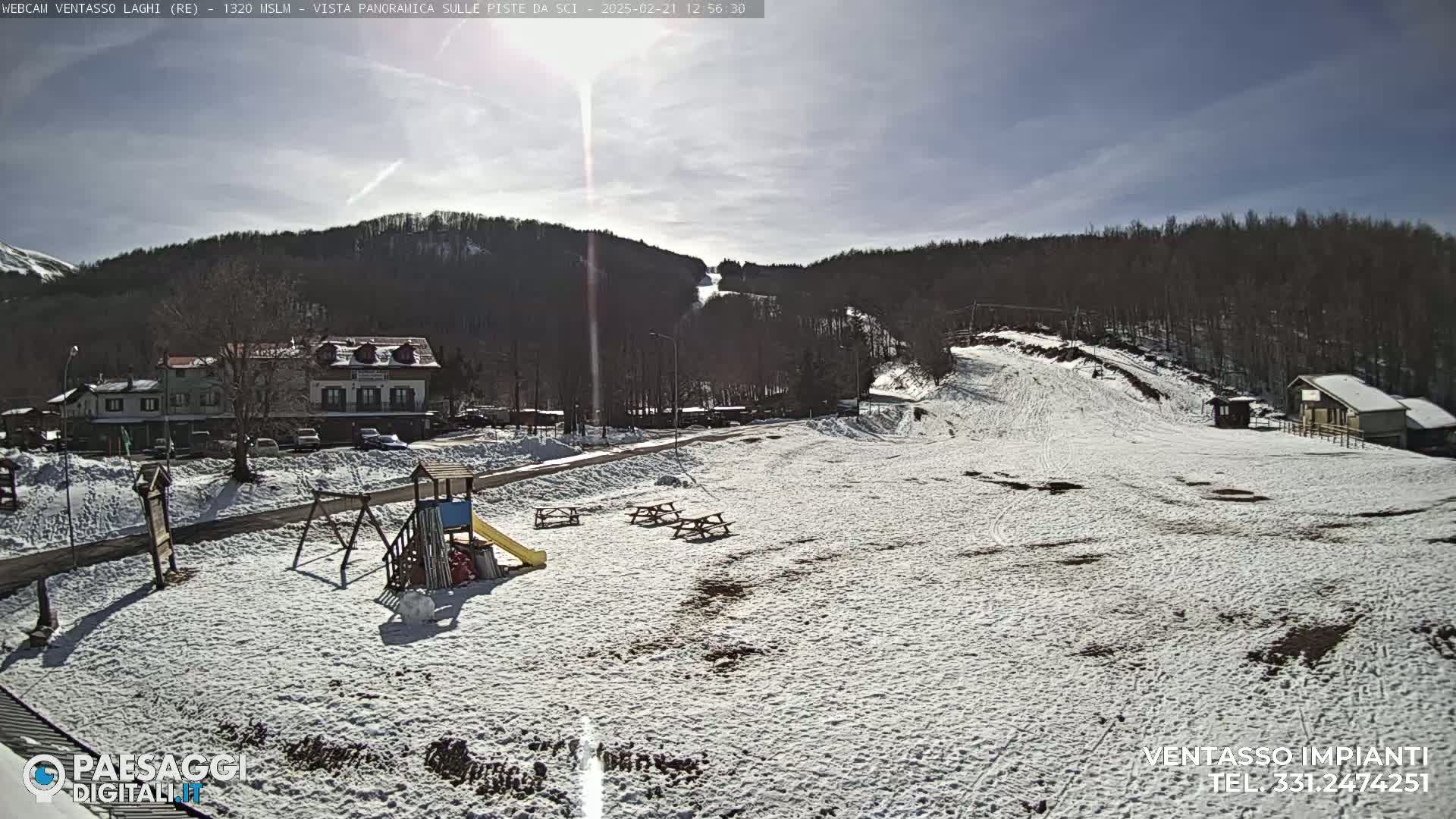 A snow-covered playground and picnic area sit below a ski slope on a sunny day, with buildings and trees visible in the background.