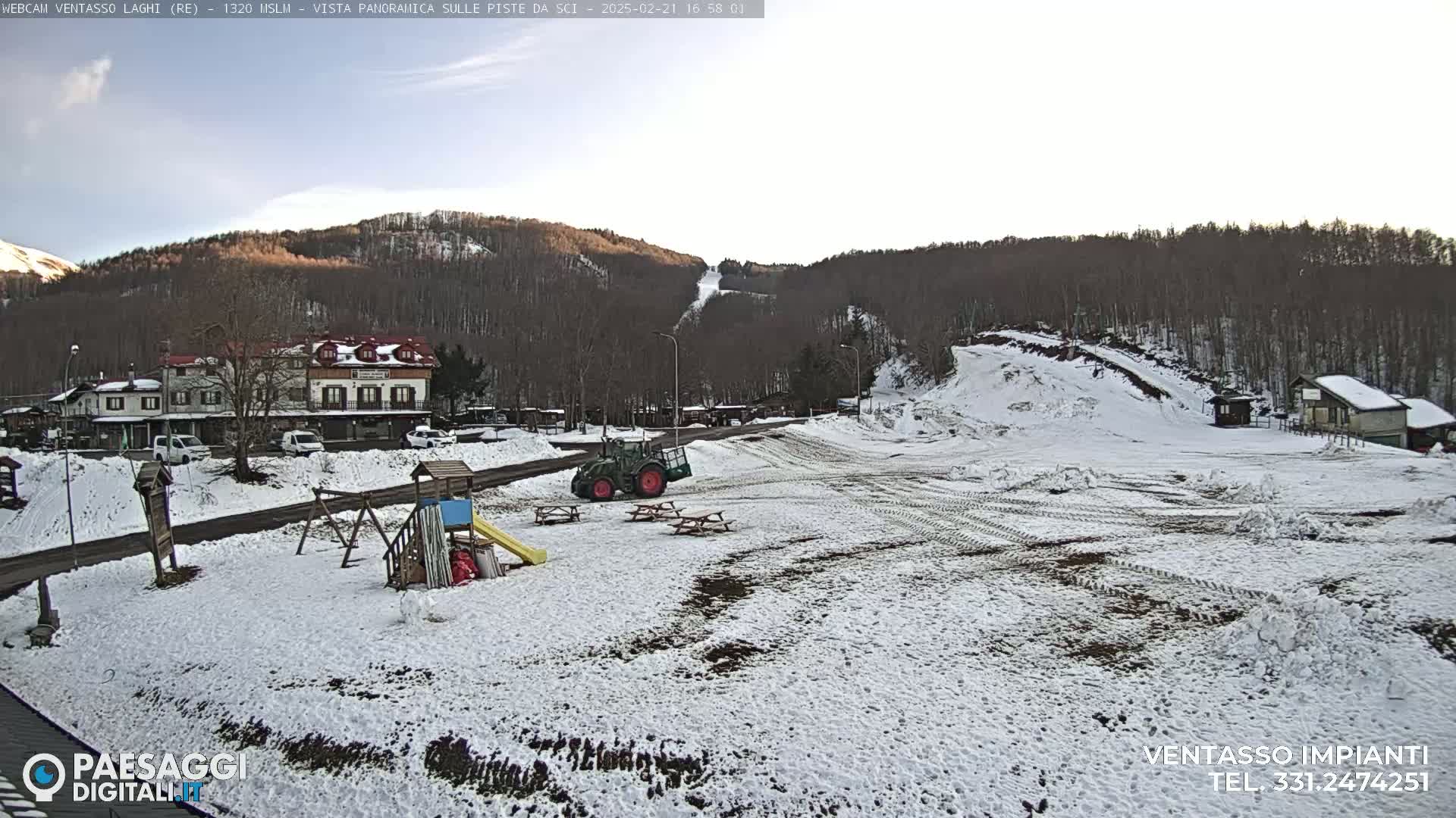 A snow-covered landscape shows a small village nestled below a ski slope on a partly cloudy day.