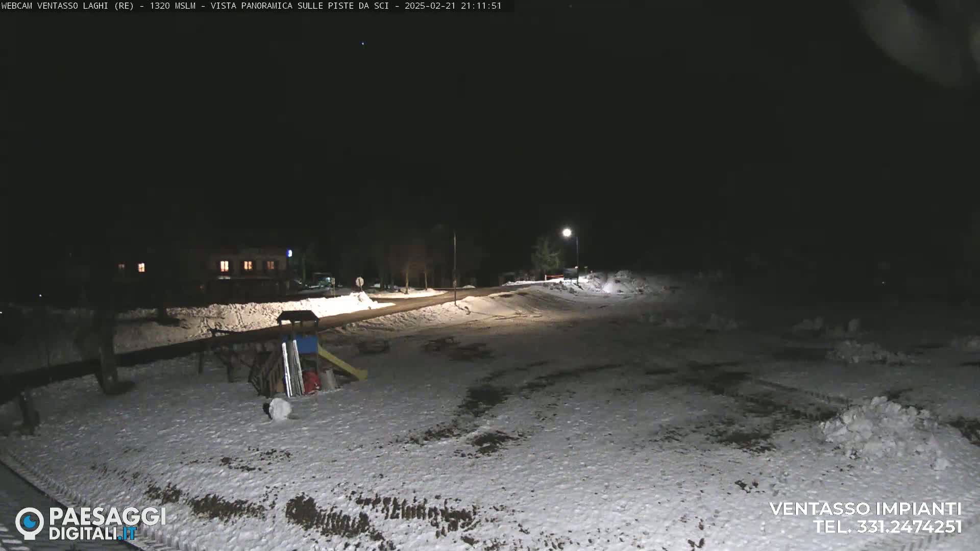 A snow-covered area at night, illuminated by a lamppost, shows a road, buildings in the distance, and some playground equipment.