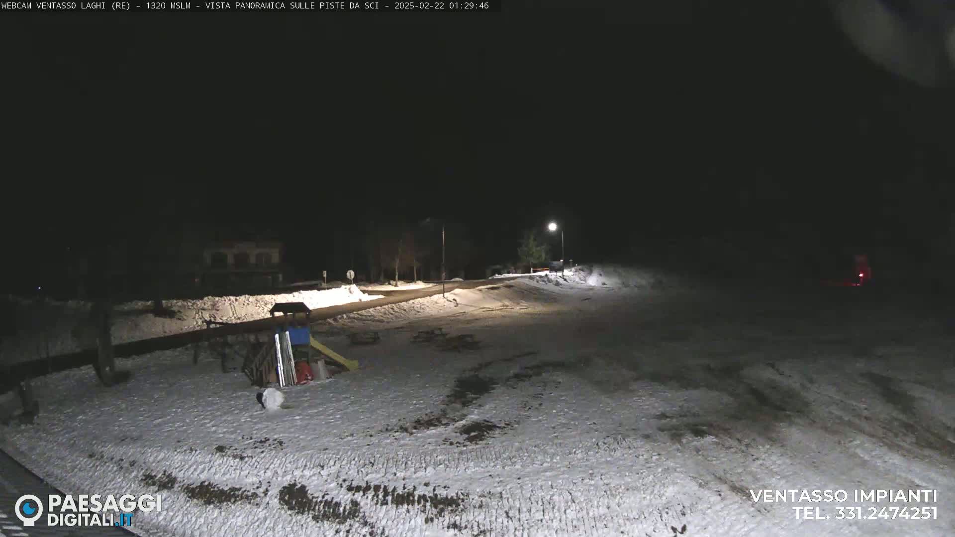 A snow-covered area at night is illuminated by a few lights, showing a road, some buildings in the distance, and playground equipment.