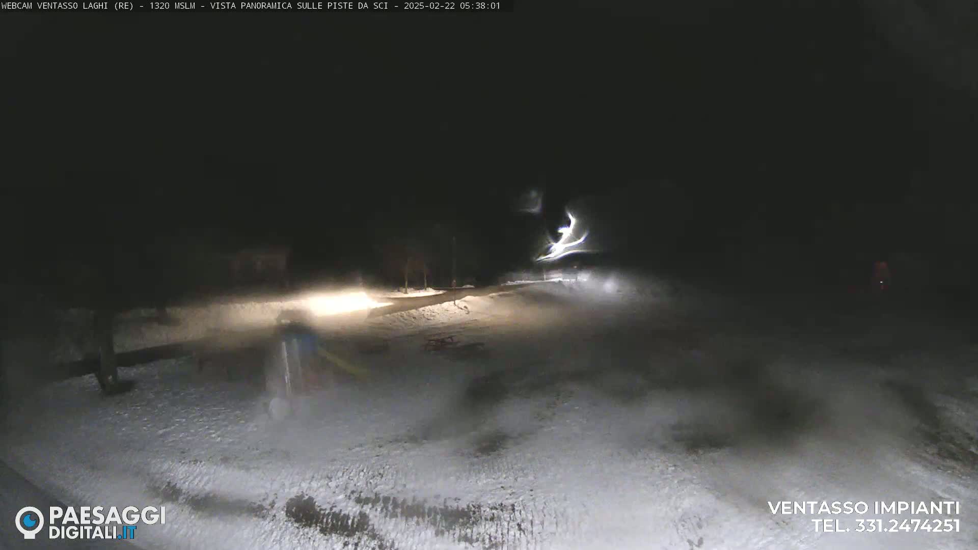 A nighttime, snow-covered scene is illuminated by vehicle headlights, showing a sparsely populated area with some trees and a road.