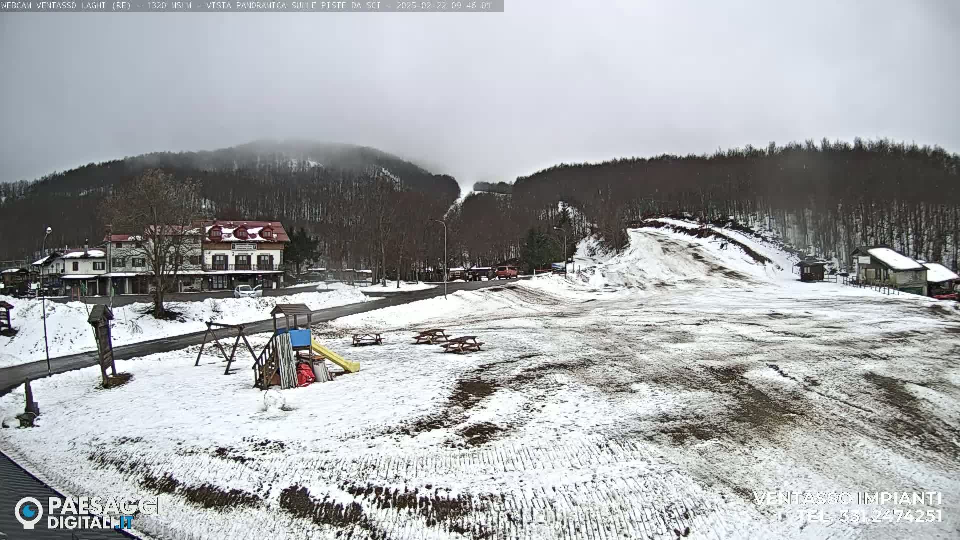 A snow-covered landscape with a building, playground equipment, picnic tables, and a ski slope under an overcast sky.