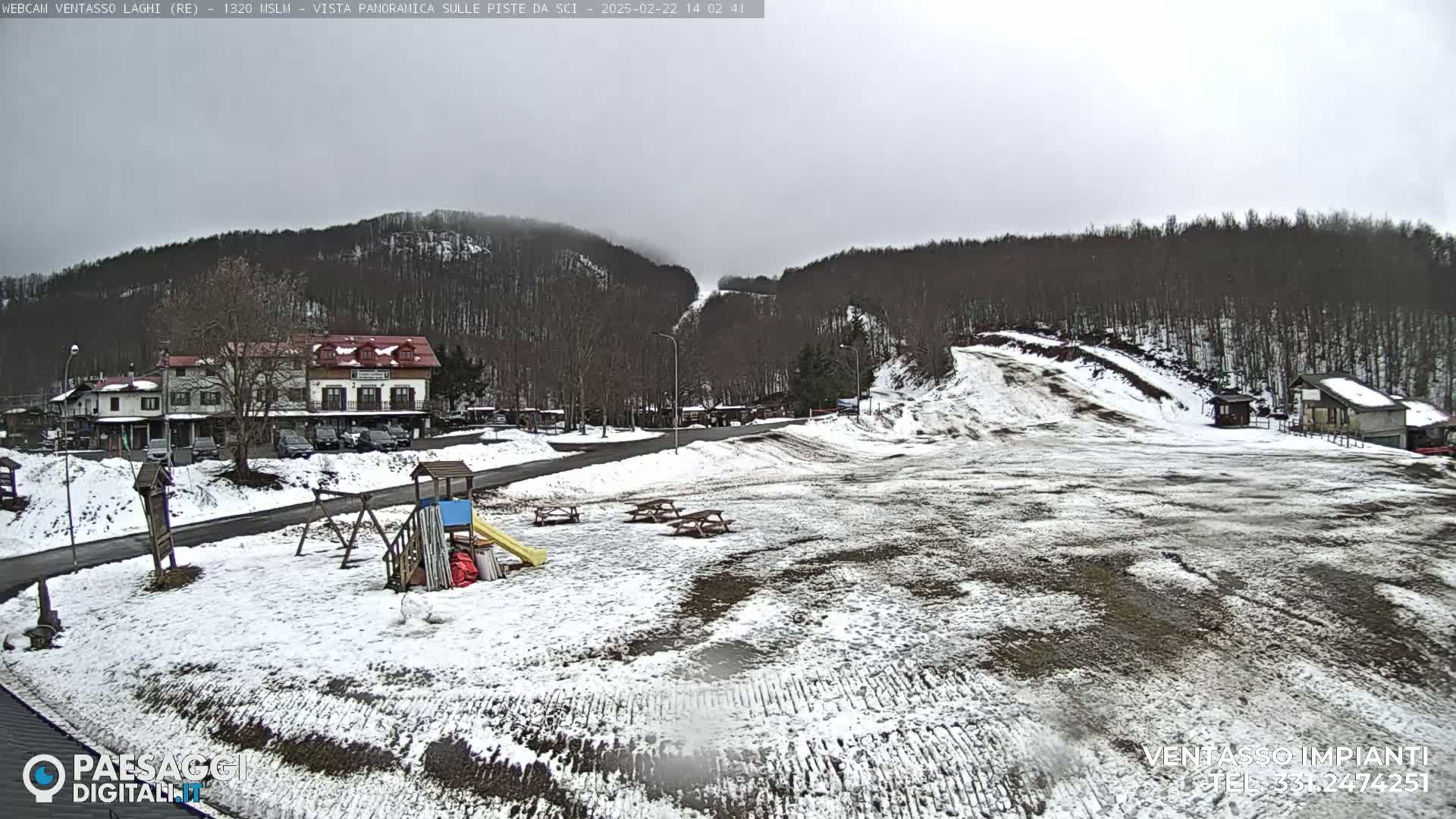 A snow-covered landscape with a few buildings, a playground, and picnic tables is visible under an overcast sky, with hills and trees in the background.