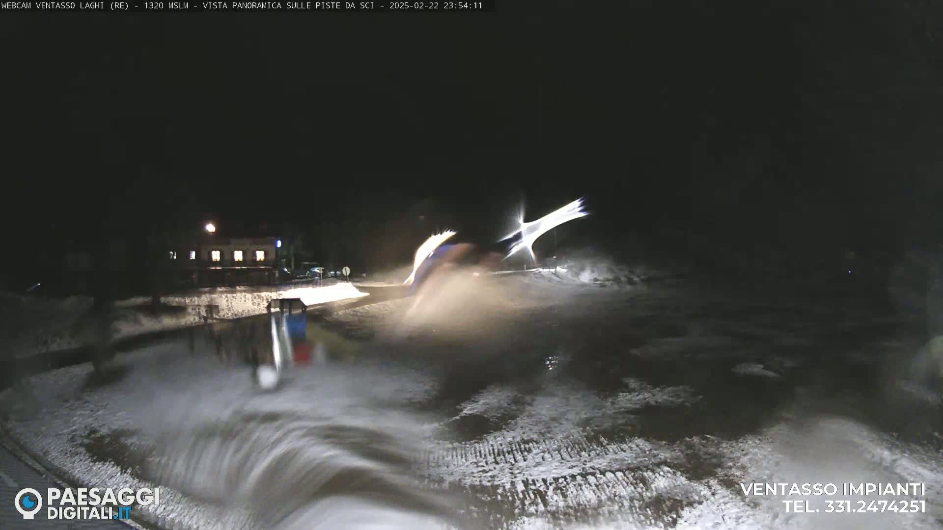 A nighttime view of a snow-covered area with a building in the background, showing some blurry motion in the foreground, possibly caused by snow or water.