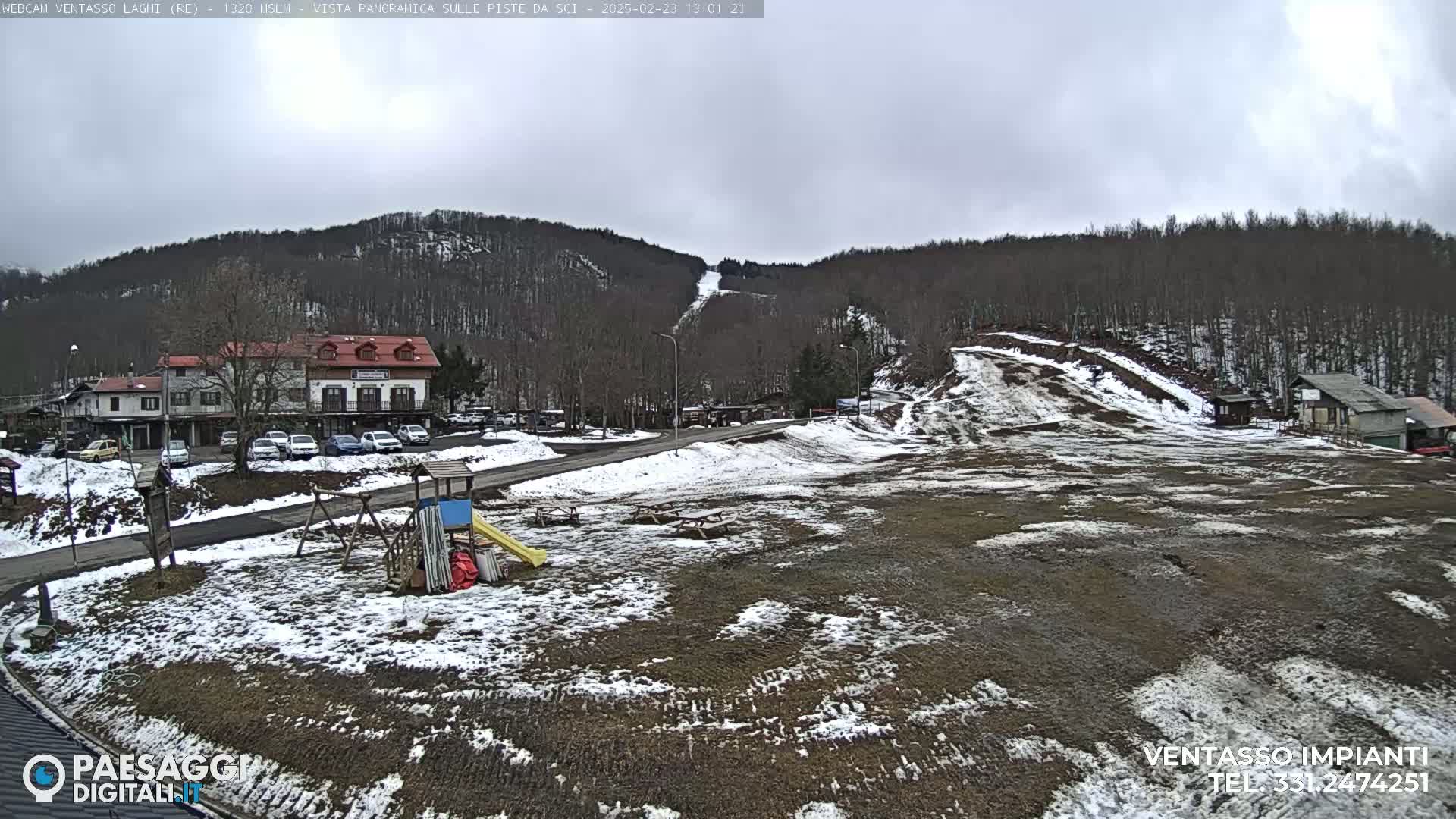 A partly cloudy day reveals a snow-dusted ski slope and surrounding village, with patches of snow on the ground and barren trees on the hillside.