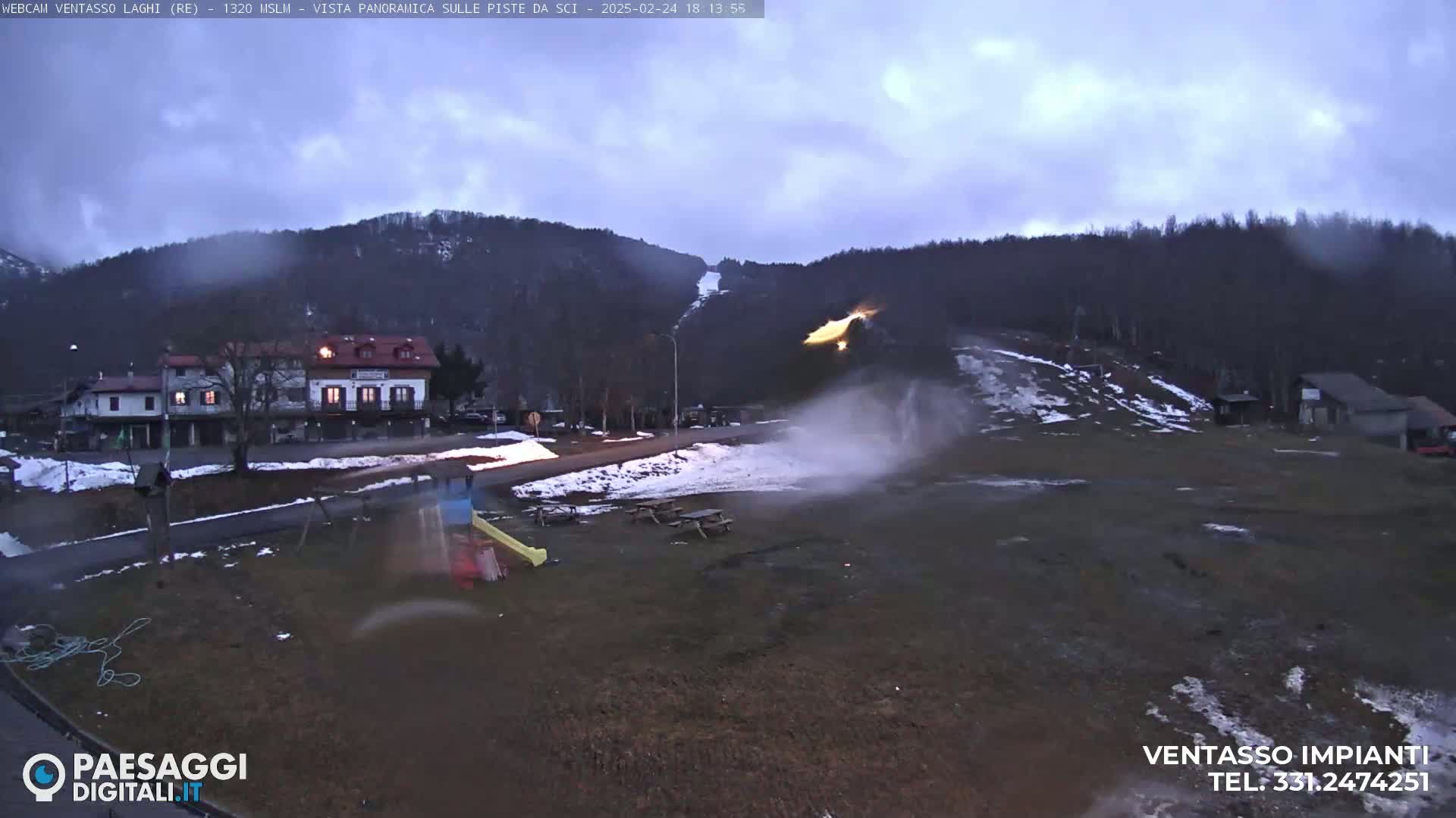 A snow-dusted landscape, under a cloudy sky, shows a small town nestled at the base of a hill with a ski slope, and a plume of snow or spray emanating from it.