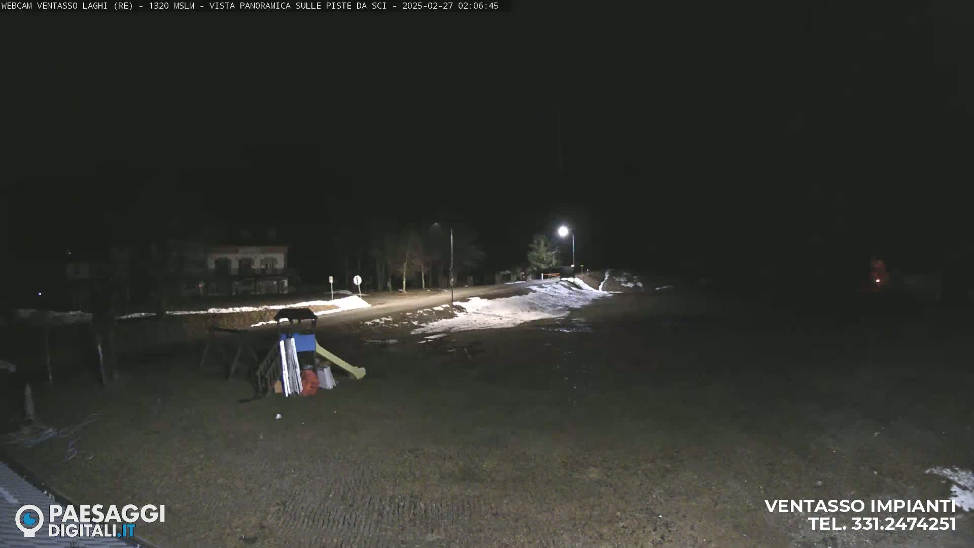 A nighttime, snow-dusted scene shows a road leading to a building, with some playground equipment and patchy snow visible in the foreground under artificial light.