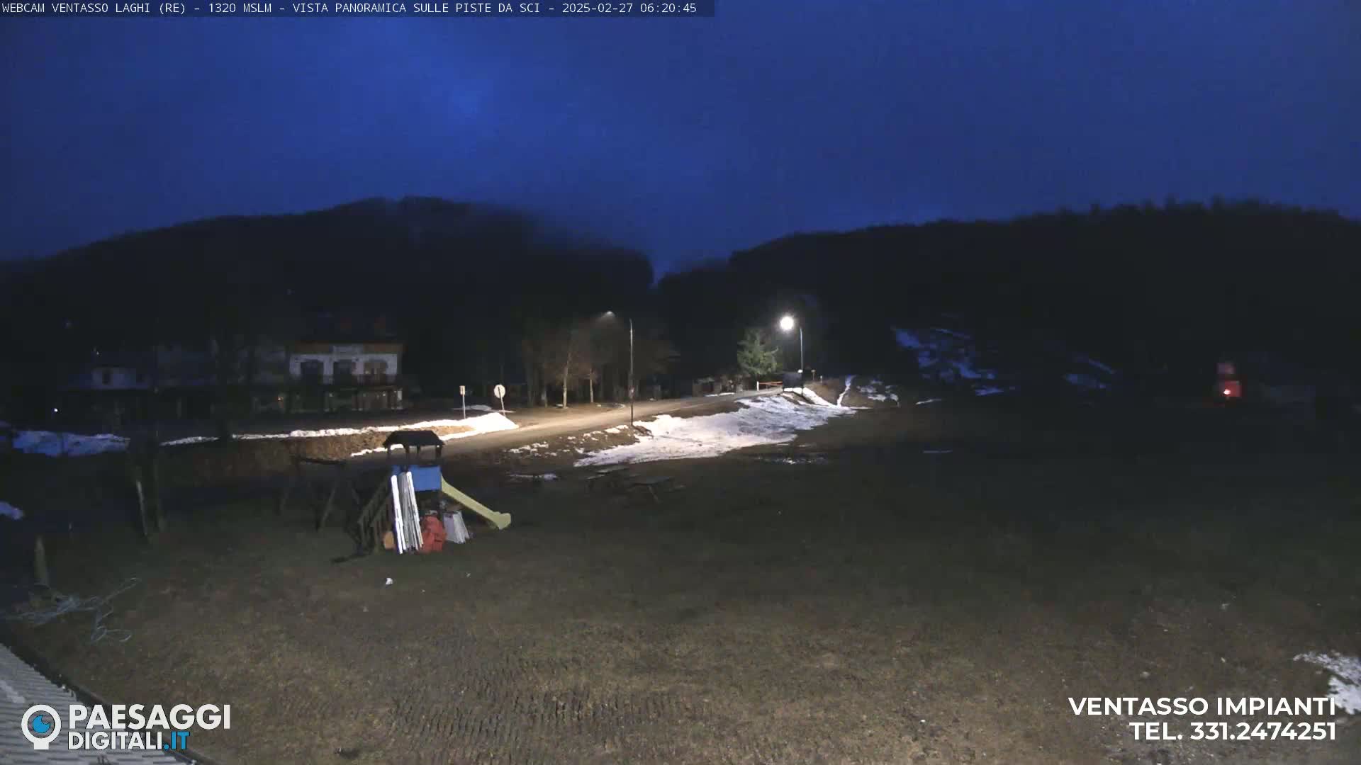 A nighttime view of a sparsely populated area with some snow on the ground, a playground, and hills in the background under a dark blue sky.