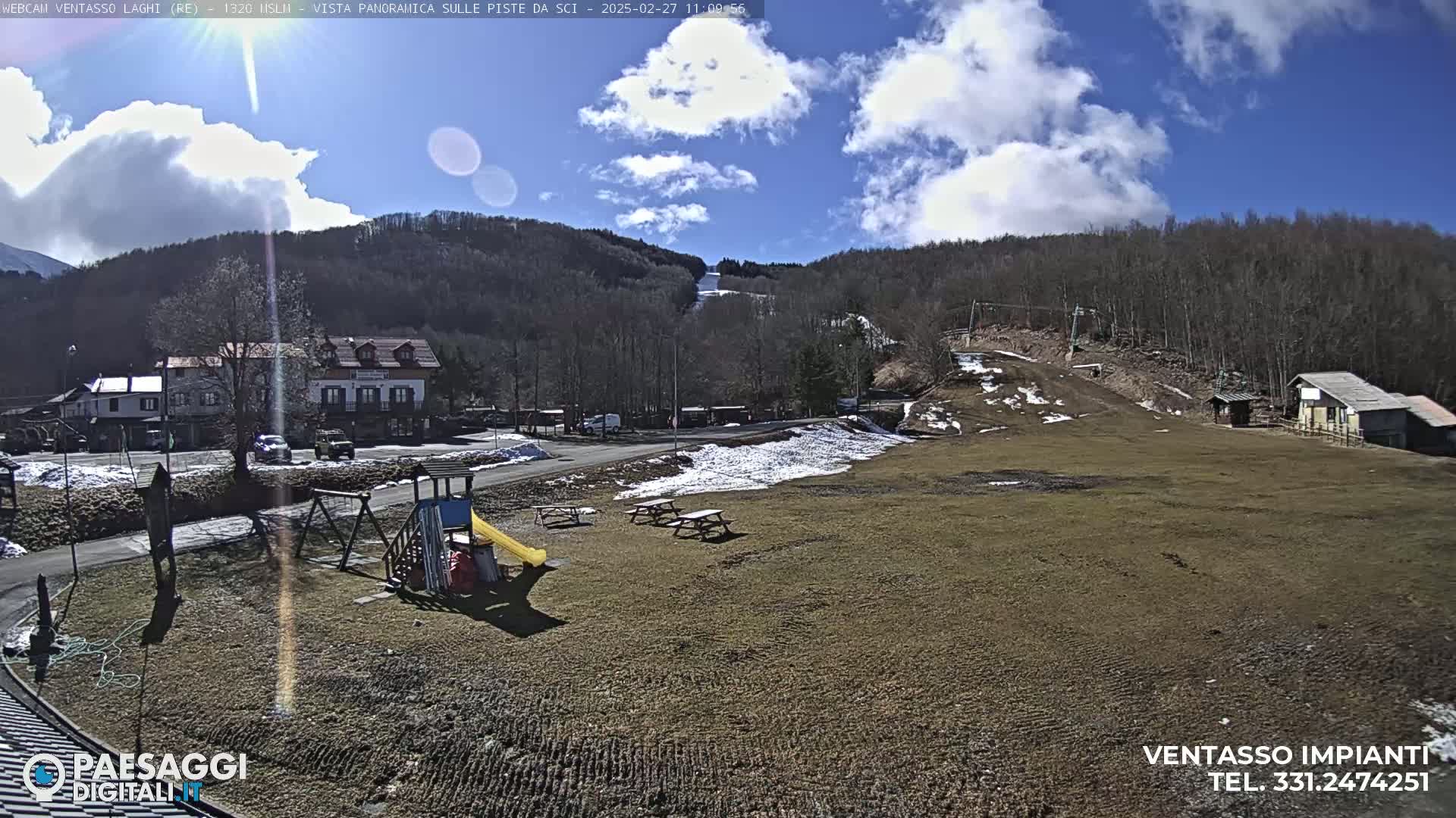 A mostly sunny day reveals a grassy area with a playground, picnic tables, and a view of a ski slope in the background, with some snow patches remaining.