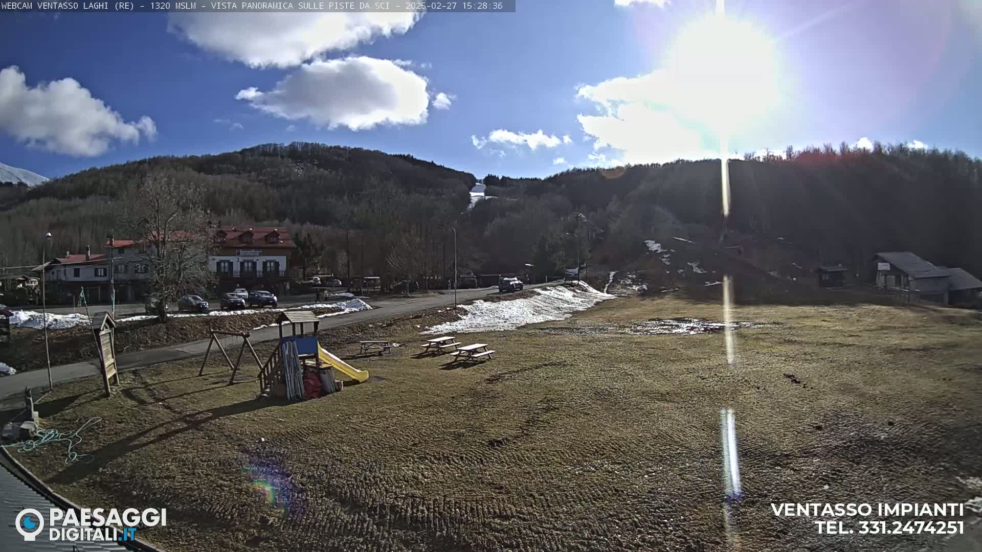 A sunny day reveals a mountainside village with a playground, picnic tables, and a ski slope in the background, with some patches of snow remaining.