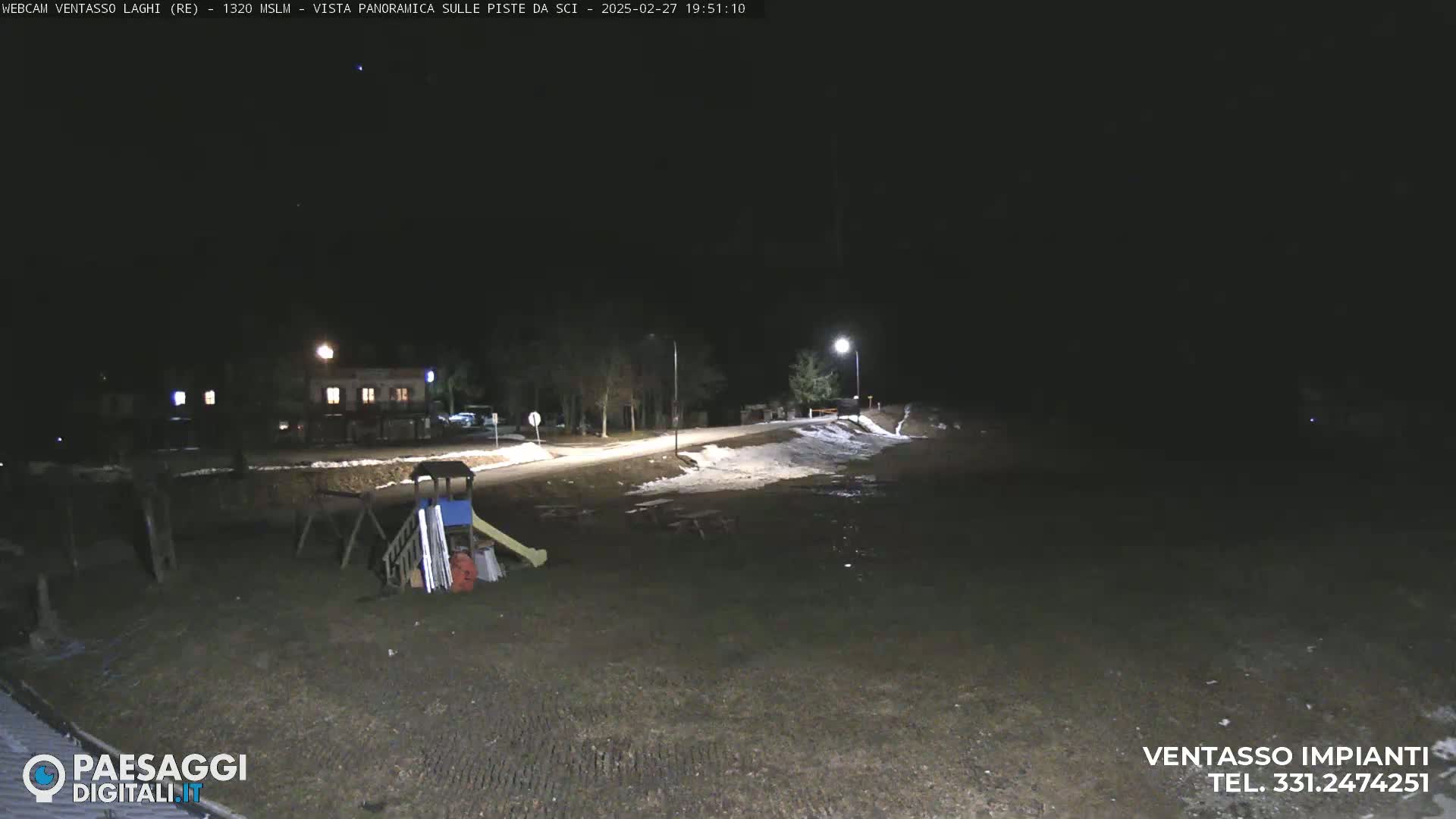 A nighttime, snow-dusted playground and road are illuminated by streetlights.