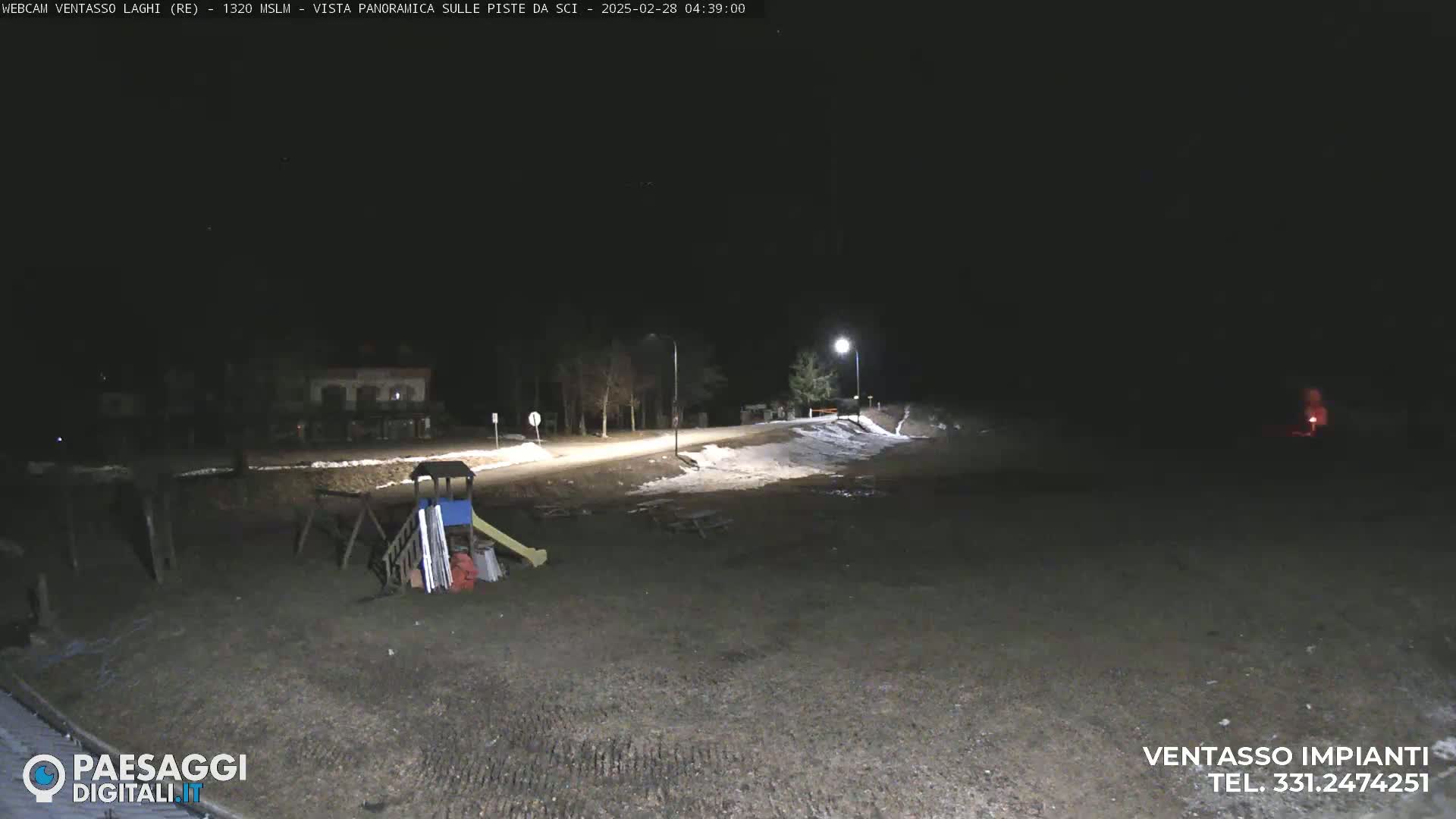 A nighttime view of a mostly empty playground near a road, with some patches of snow on the ground and a few lights illuminating the scene.