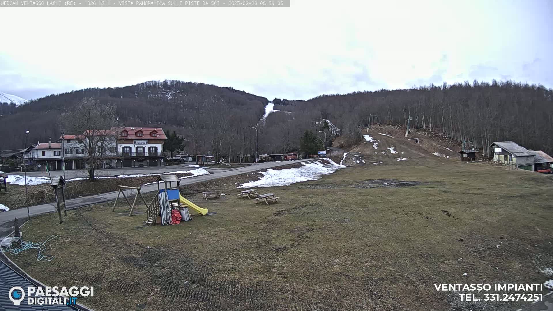 A playground with a slide and swings sits near a small town nestled in a valley, with a ski slope visible on the hillside under a cloudy sky.