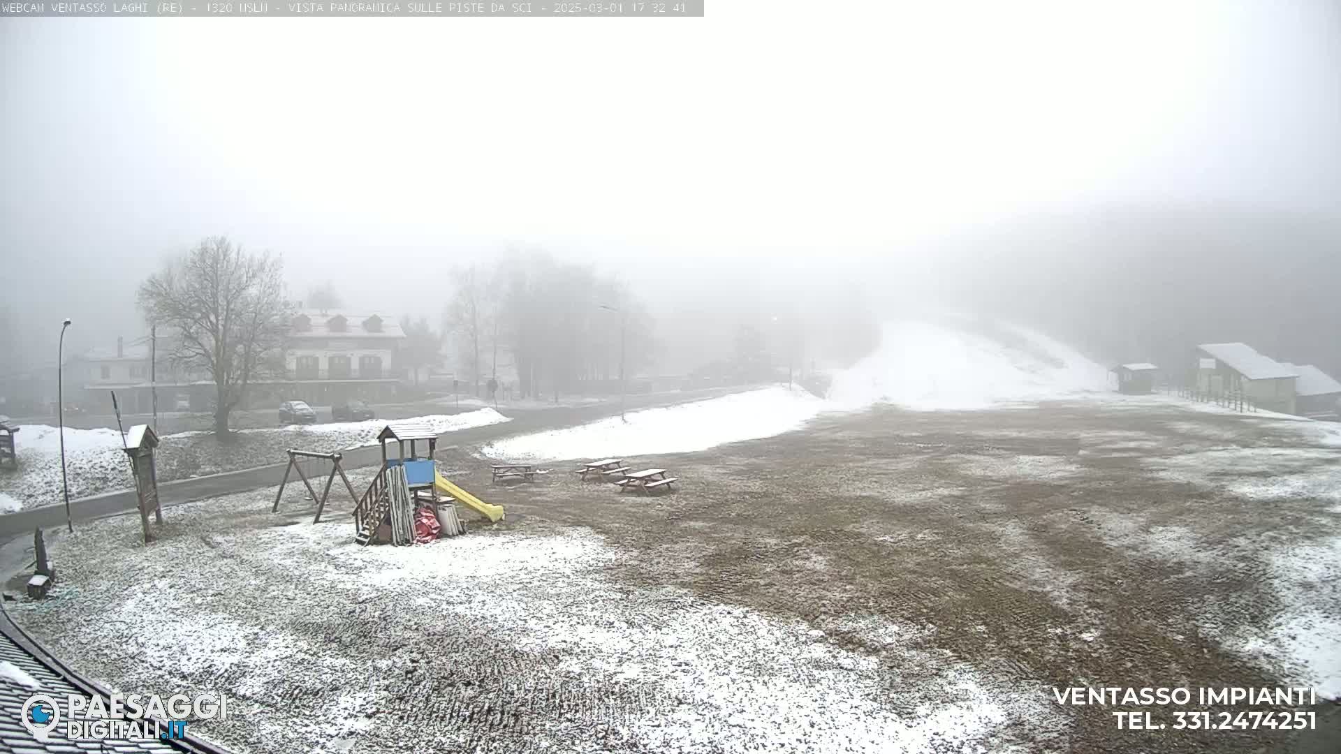 A foggy, snowy landscape shows a playground, picnic tables, and buildings in the distance.