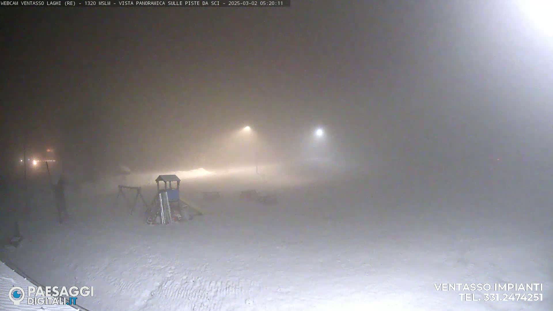 A snow-covered playground is dimly lit by two distant lights in heavy fog or blowing snow at night.