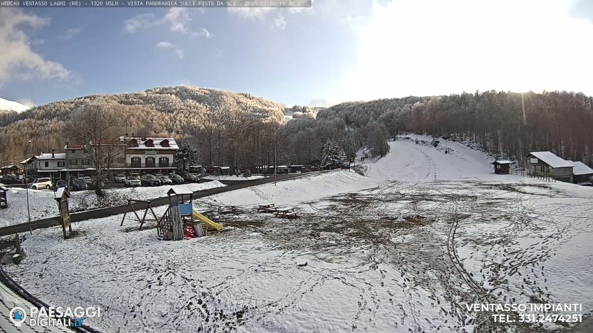 A snow-covered playground and ski slope are situated below a line of snow-dusted trees and mountains under a mostly sunny sky.