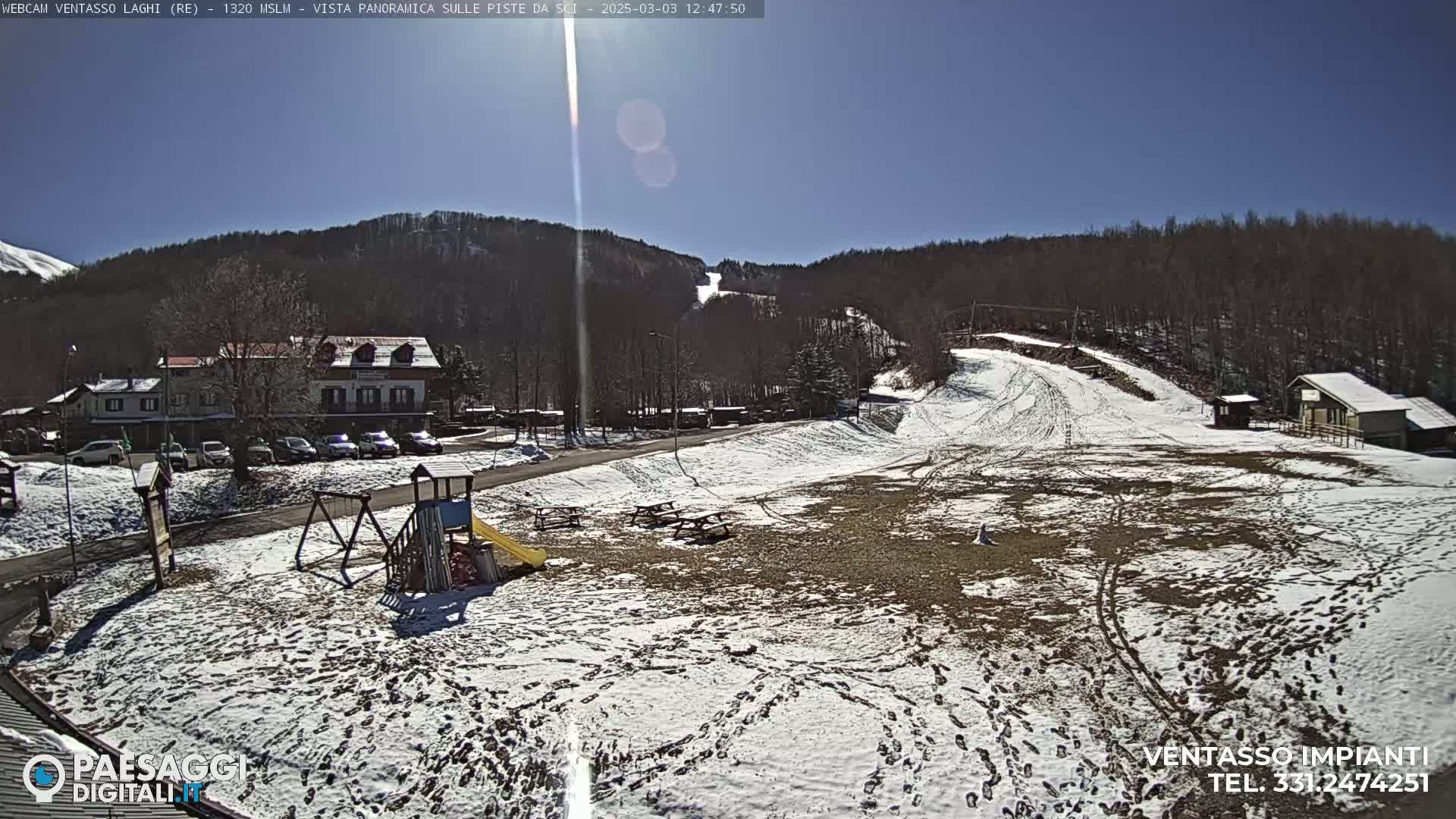 A sunny, snow-dusted ski area with a playground, buildings, and ski lifts visible against a backdrop of leafless trees.