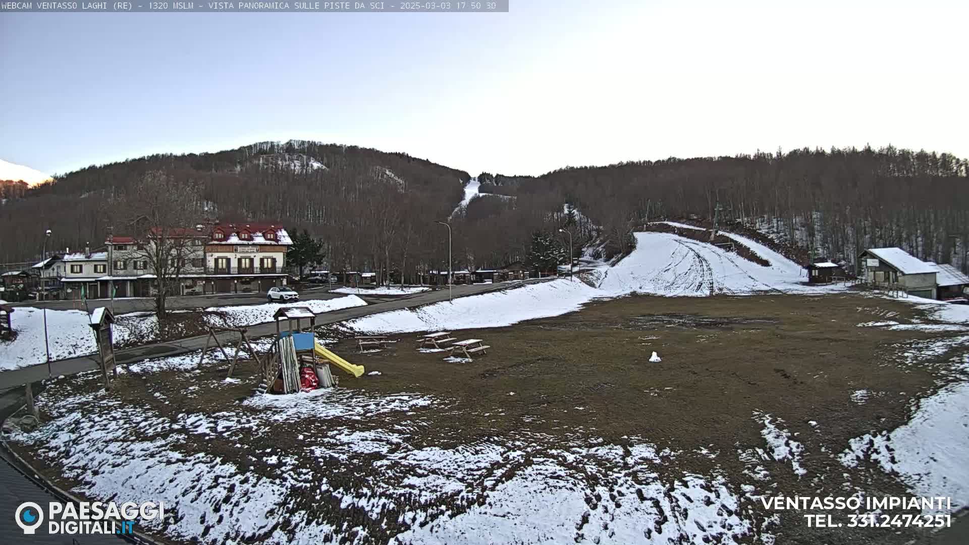 A mostly clear day shows a snow-dusted ski resort area with a building, playground equipment, picnic tables, and ski slopes.