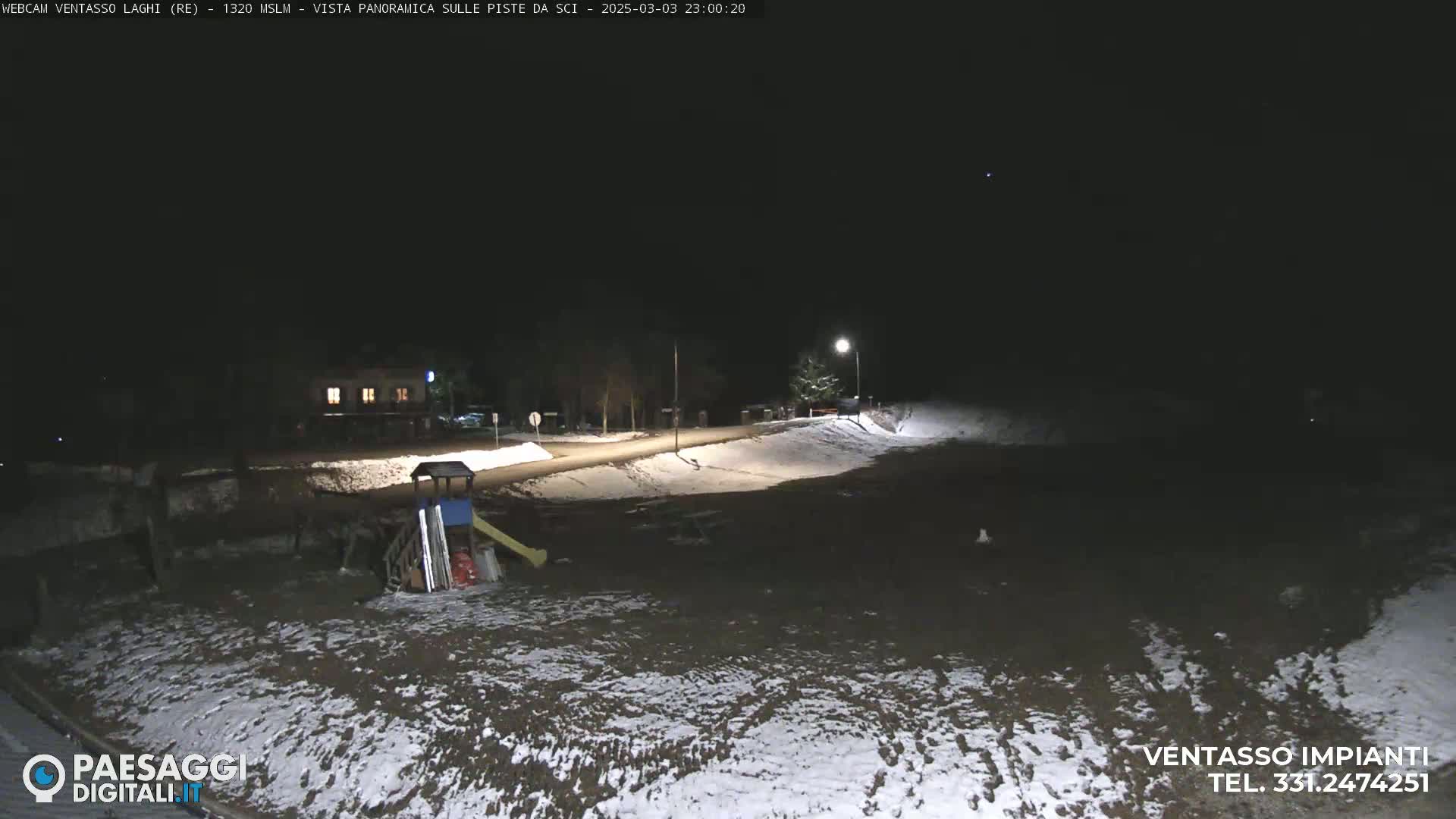 A nighttime, snow-dusted, outdoor scene shows a playground near a road and a building under a dark sky.