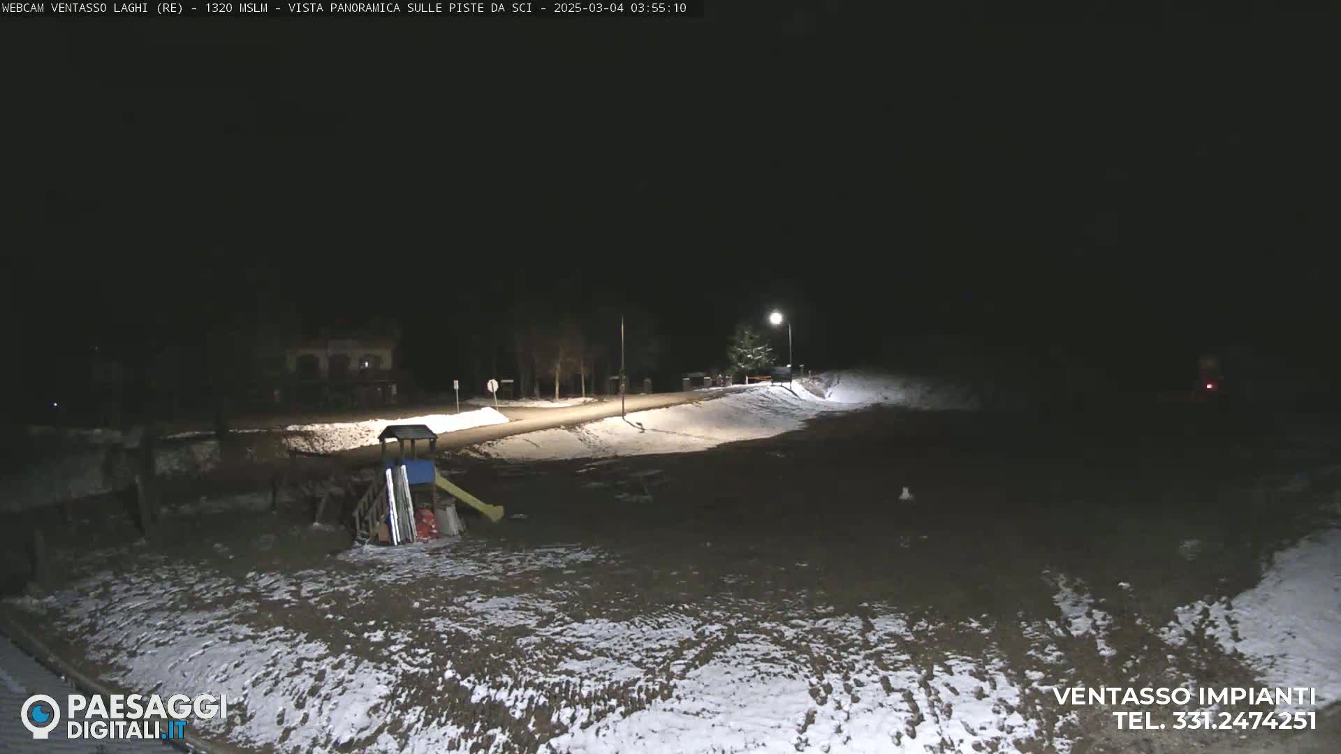 A nighttime, snow-dusted, outdoor scene shows a playground and a road lit by a streetlamp.